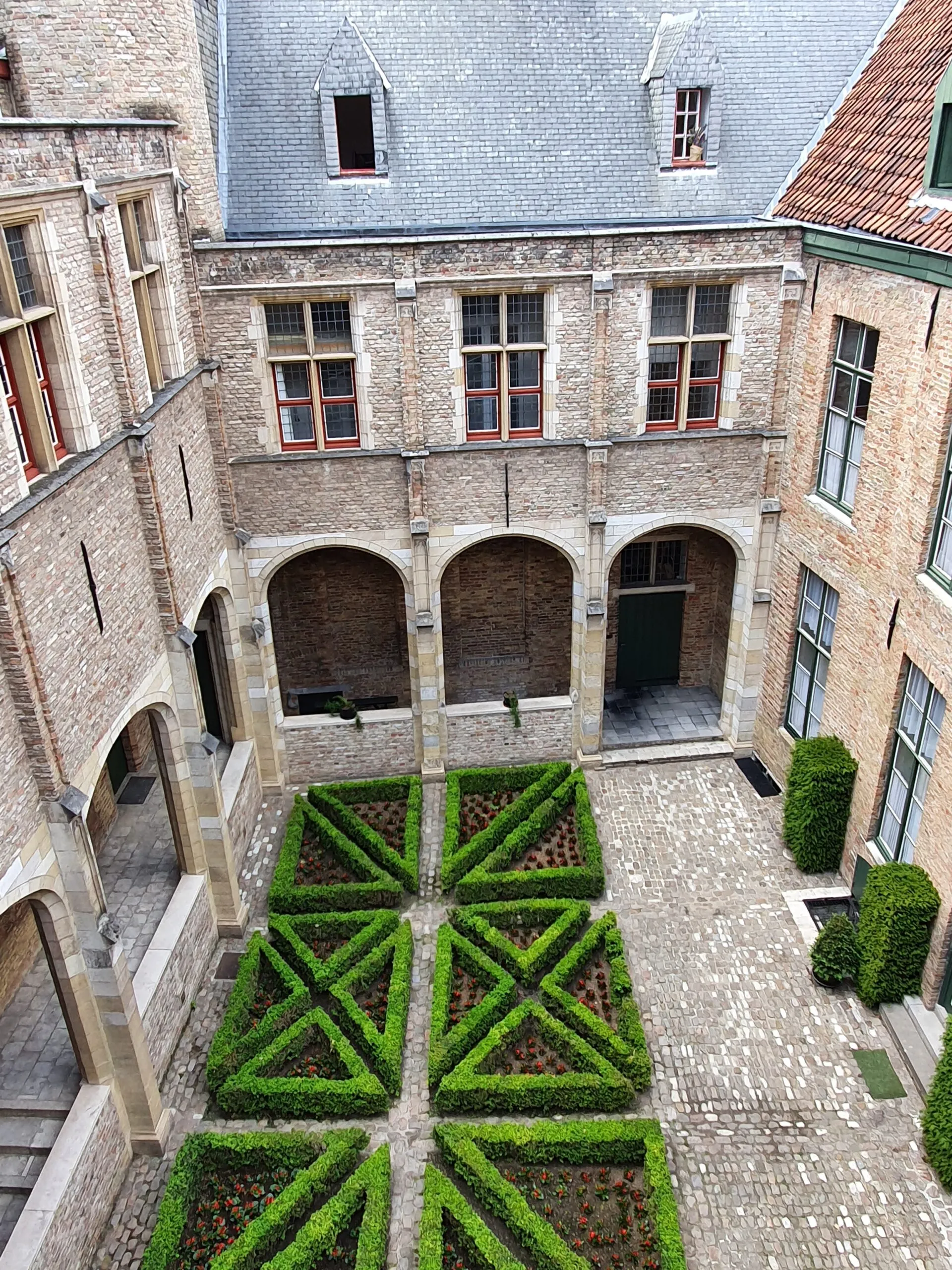 Vista aérea de un patio cerrado con un jardín de setos geométricos, pasillos de ladrillo con arcos, pavimento de adoquines y un edificio histórico con tejado de pizarra en Hof Bladelin.
