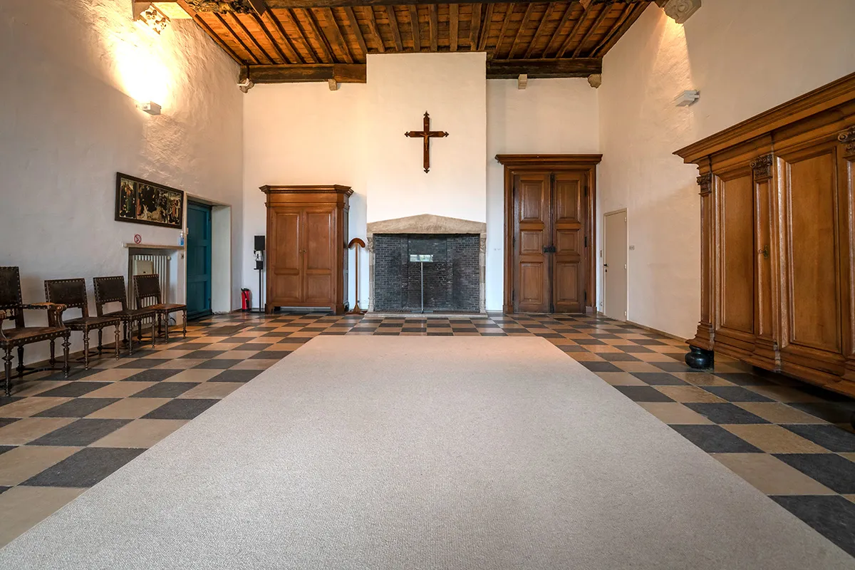 Interior de un salón histórico en Hof Bladelin con suelo de cuadros blancos y negros, una alfombra larga de color blanco, vigas de madera en el techo y una cruz en la pared del fondo.