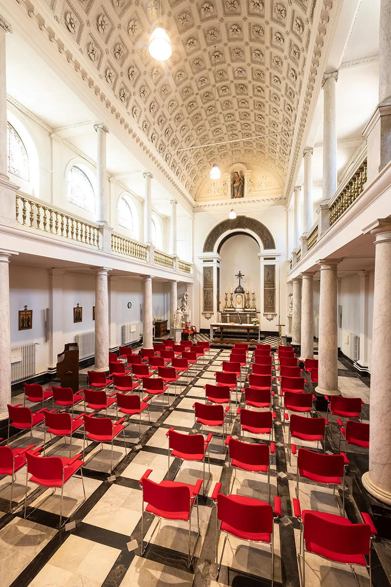 Interior de una capilla con filas de sillas rojas orientadas hacia un altar, un techo abovedado ornamentado, columnas blancas y un suelo a cuadros