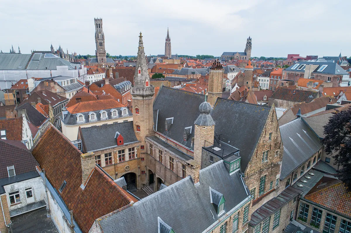 Vista aérea del Hof Bladelin que muestra edificios históricos de ladrillo con tejados rojos y grises y pequeñas torres, con el horizonte de la ciudad de Brujas y el Campanario al fondo.