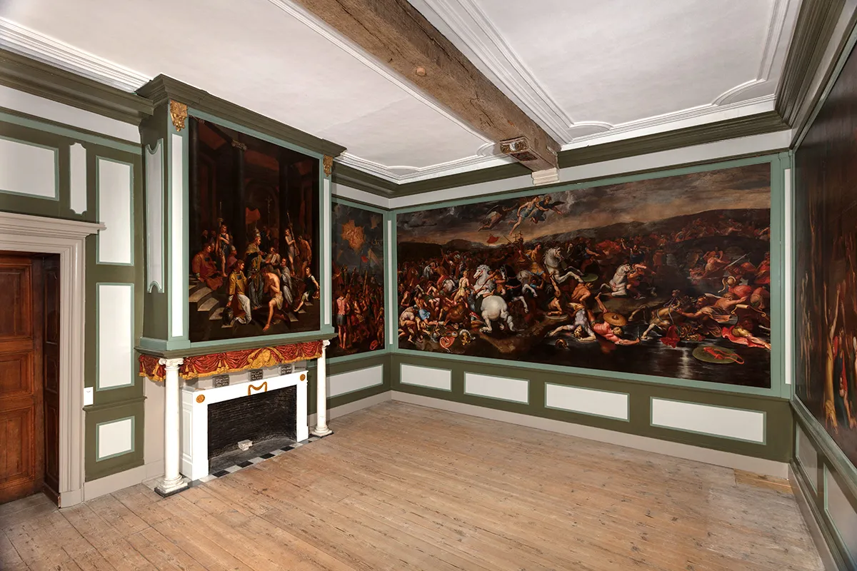 Historic room at Hof Bladelin with large narrative paintings covering the walls, a white fireplace with decorative mantel, wooden floor, and exposed ceiling beams