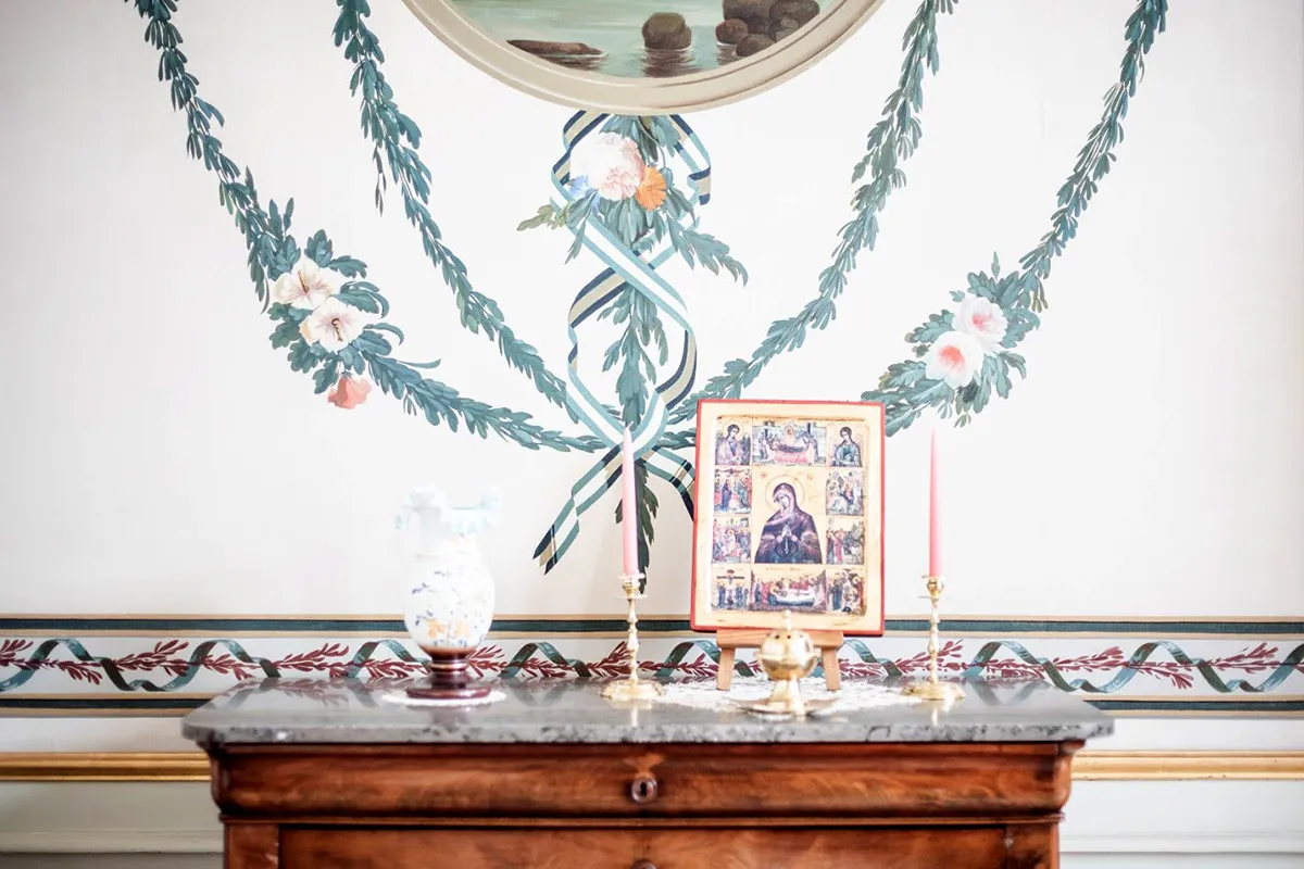 A wooden console table with a marble top holding a white vase, brass candlesticks with pink candles, and a framed religious icon, set beneath a wall painted with decorative green garland wreaths and floral ribbon motifs.