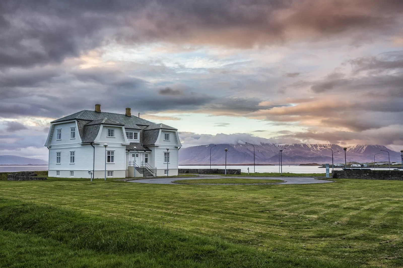 White two-story house with a green lawn in front, waterfront and snow-capped mountains in the background under a dramatic cloudy sky