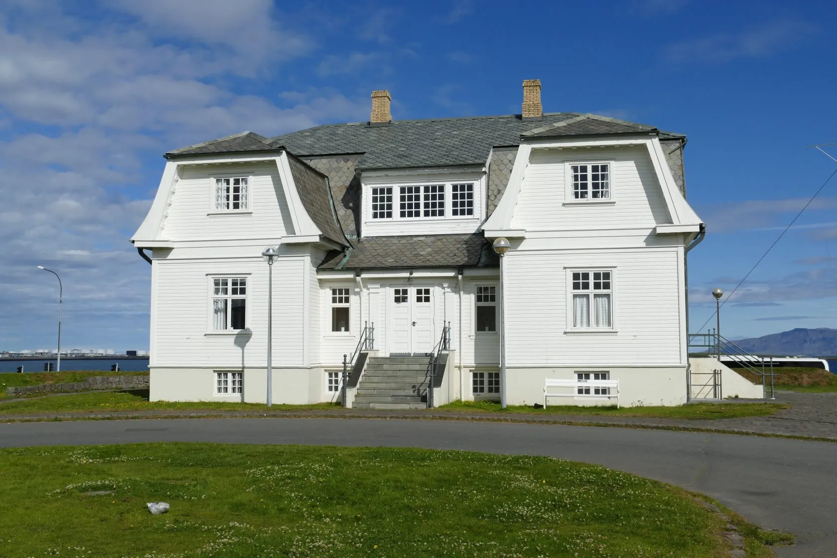 White two-story historic house with a gray mansard roof, two chimneys, and a central staircase entrance, set against a blue sky with green lawn in the foreground
