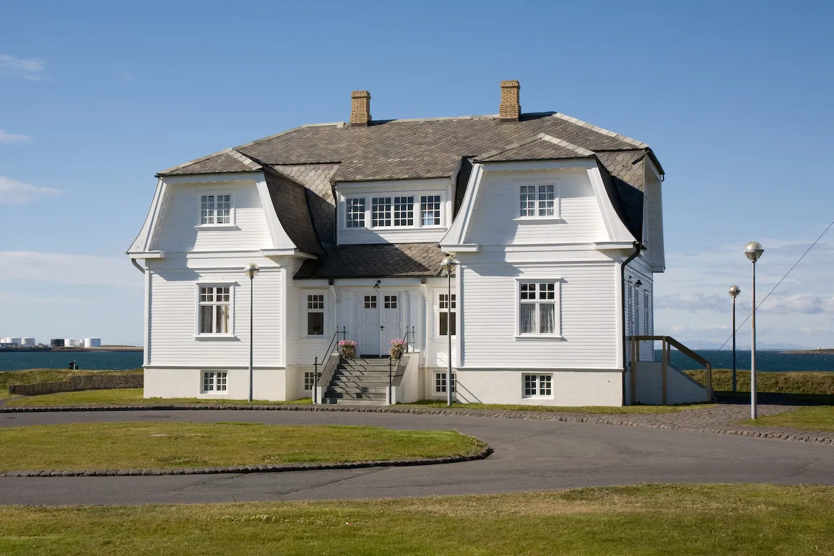 A white two-story wooden house with a gray shingled roof, two chimneys, and a central staircase entrance, set against a blue sky with water visible in the background.
