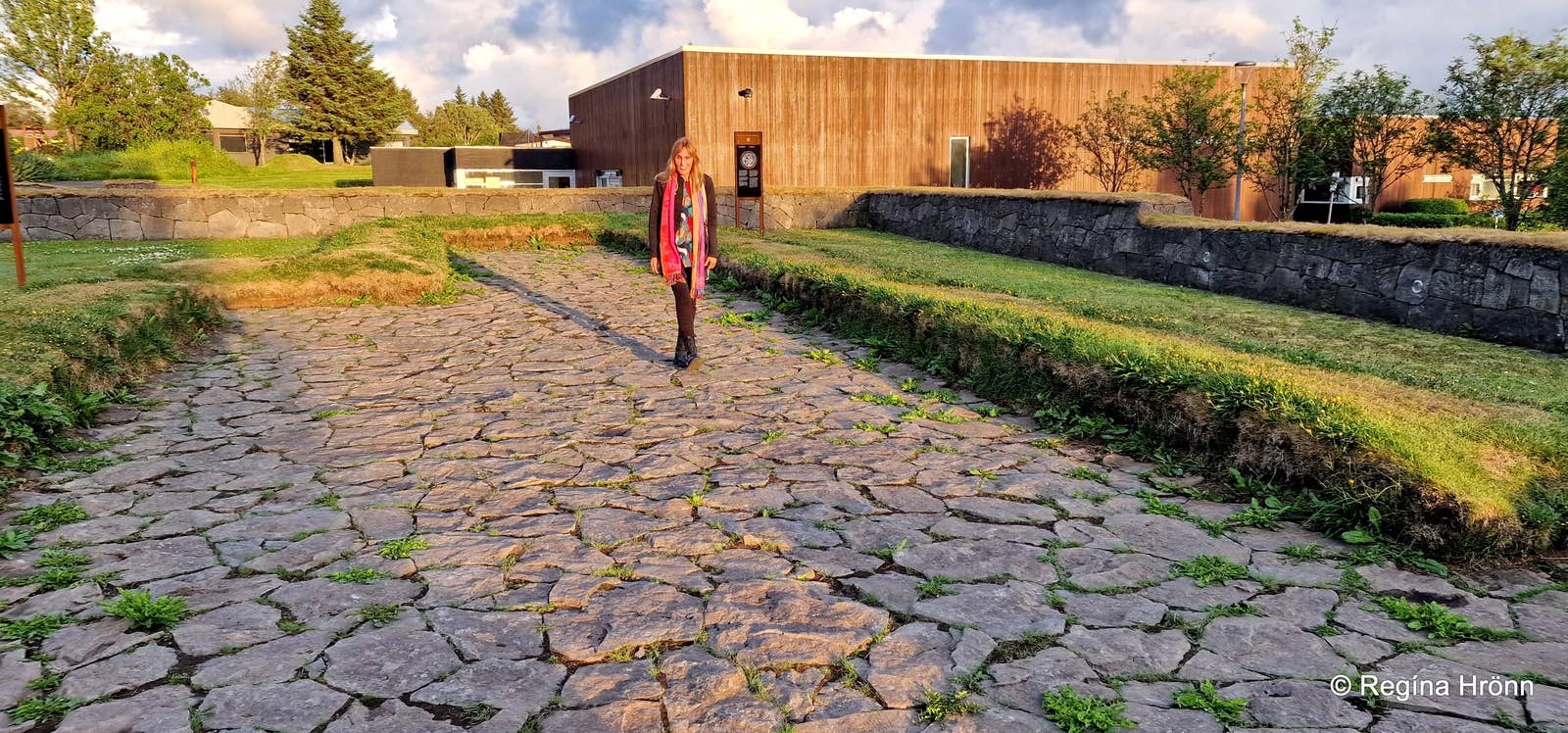 Sendero empedrado que atraviesa las ruinas de casas comunales vikingas cubiertas de hierba, con una persona de pie en el centro y un edificio moderno de madera al fondo