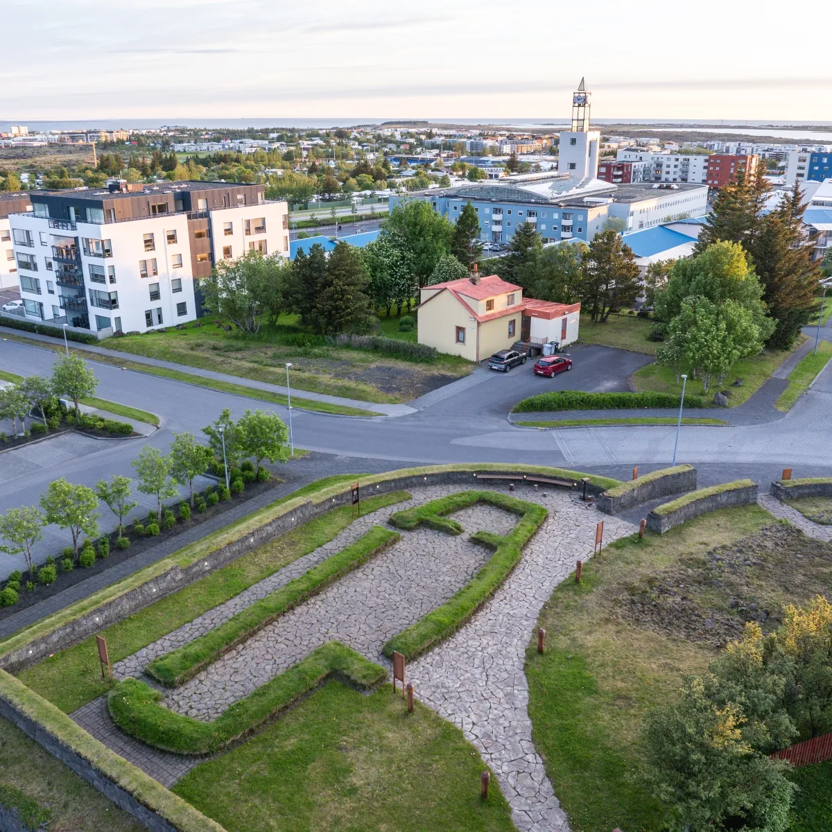 Vista aérea de los cimientos de la casa comunal vikinga de Hofstaðir, delimitados con setos y senderos de grava, con un pueblo moderno y una iglesia al fondo.