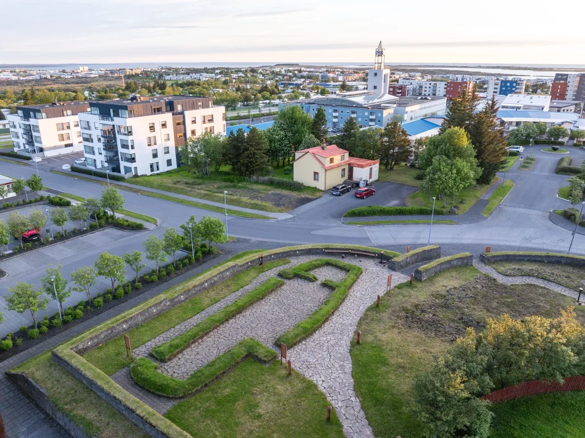 Vista aérea de los cimientos de piedra y grava de una casa larga vikinga en Hofstaðir, con edificios residenciales modernos y la torre de una iglesia visibles al fondo.