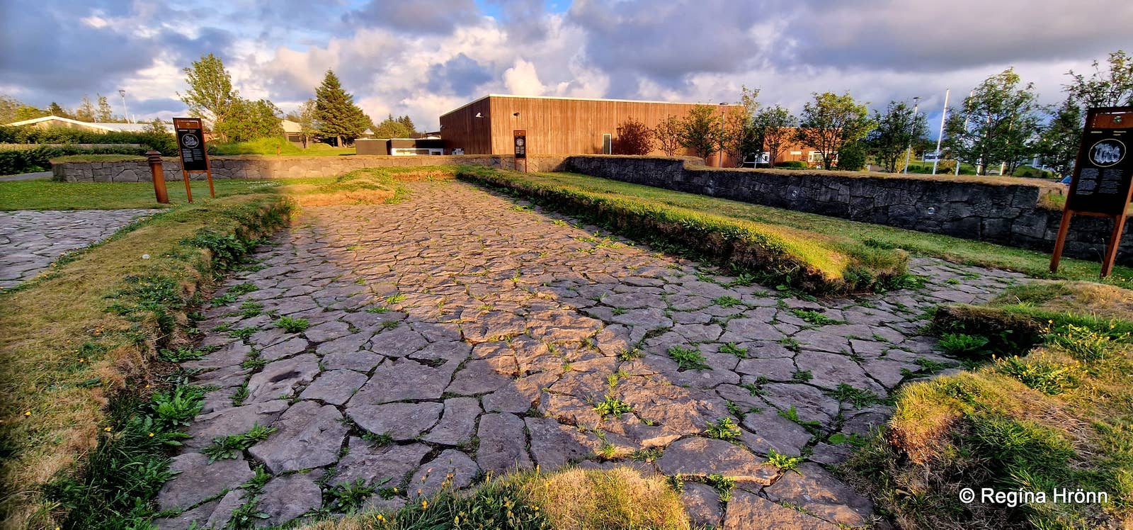Un ancho camino de adoquines conduce hacia un edificio de madera moderno, flanqueado por muros de piedra bajos y paneles informativos bajo un cielo nublado.