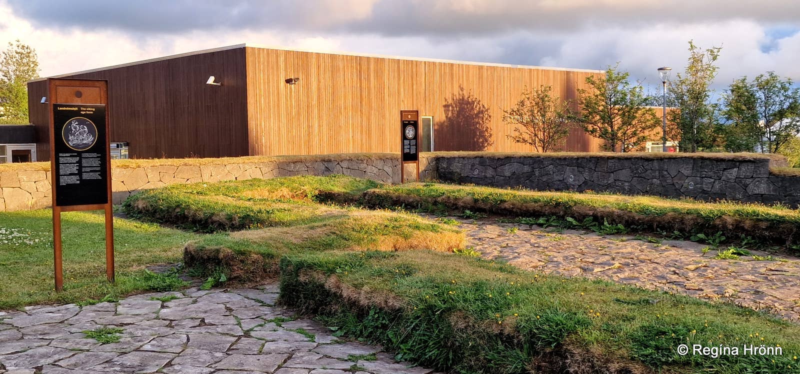 Yacimiento arqueológico al aire libre con ruinas de cimientos de piedra cubiertos de hierba de una casa comunal vikinga y un moderno edificio de exposiciones revestido de madera, con un panel interpretativo en primer plano