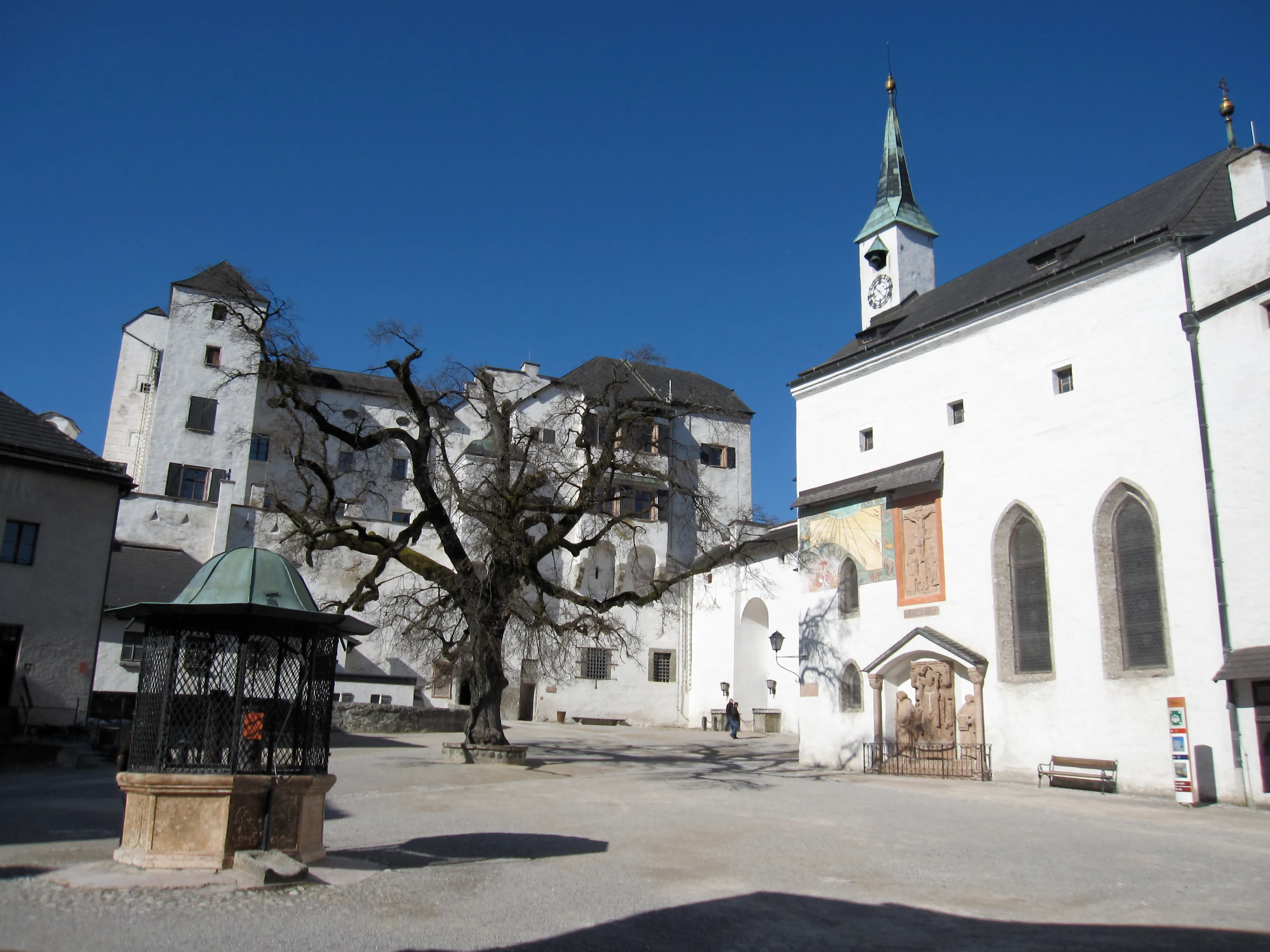 Patio interior de la fortaleza Hohensalzburg con edificios blancos, una iglesia con una aguja verde, un gran árbol desnudo y una estructura de pozo bajo un cielo azul.