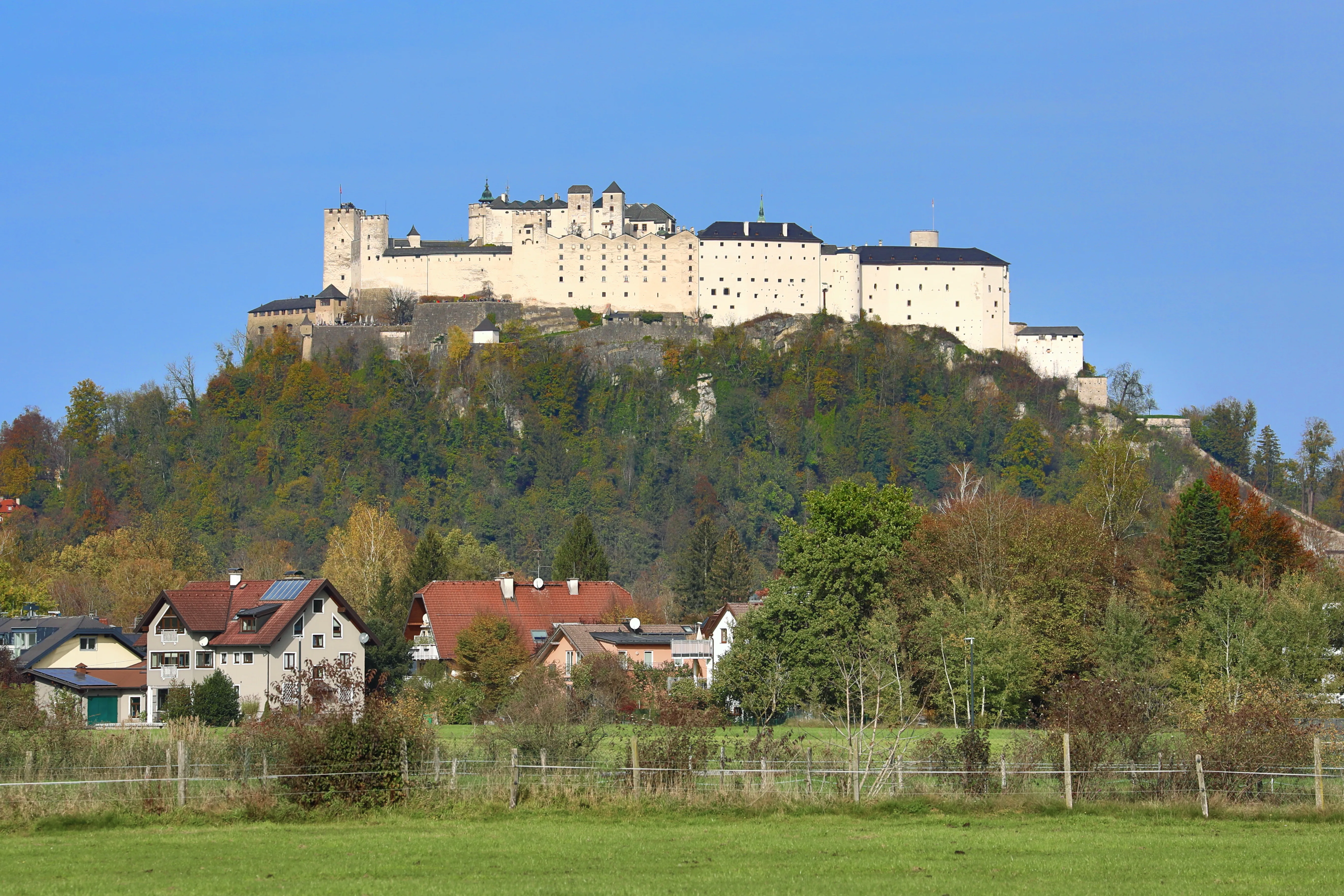 La blanca fortaleza medieval de Hohensalzburg situada sobre una colina boscosa, encima de casas y campos verdes bajo un cielo azul despejado.