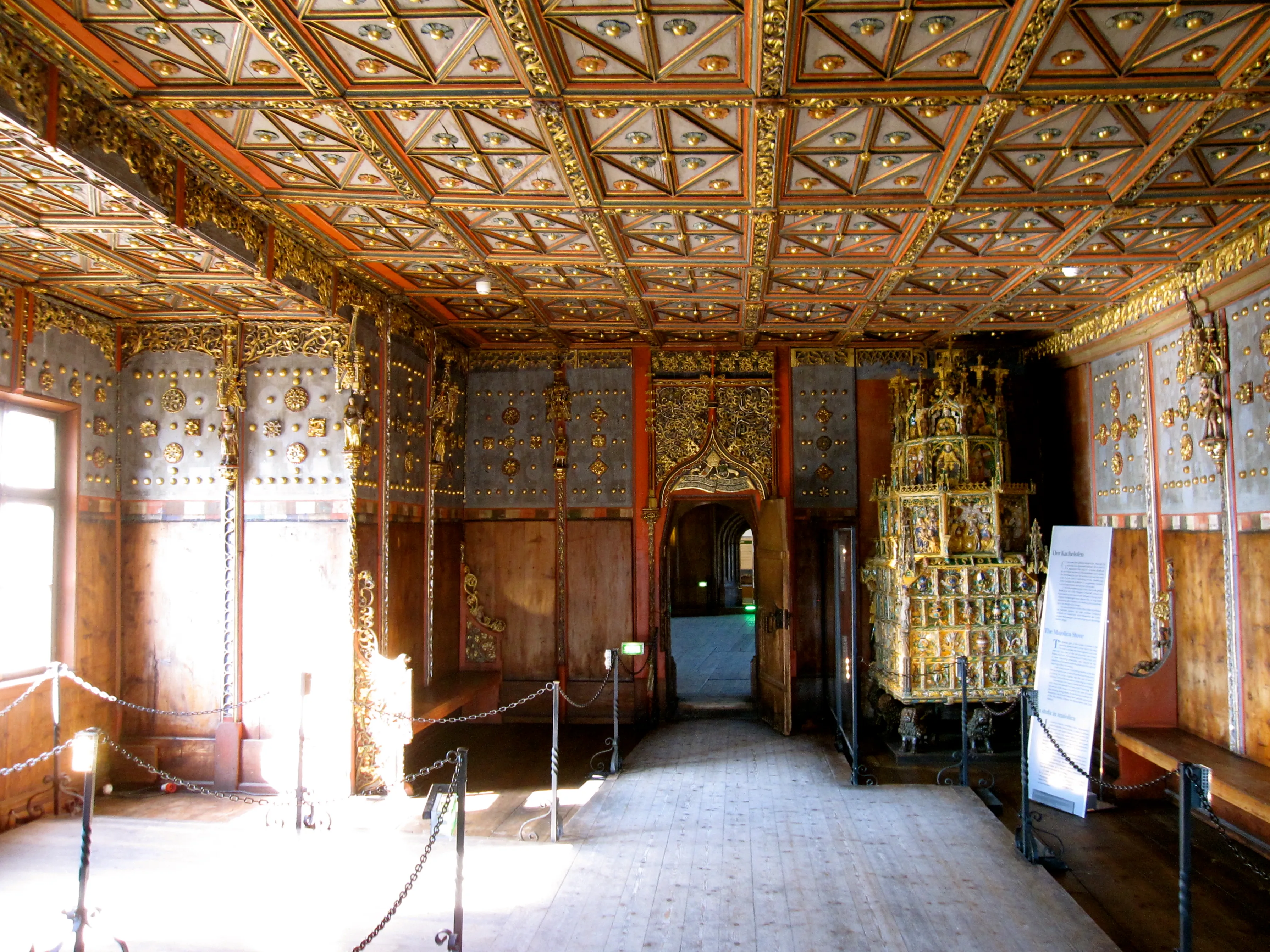 Habitación interior ornamentada con un techo artesonado de madera dorada, paneles de pared pintados con detalles en oro, una gran estructura decorativa dorada a la derecha y barreras de cuerda que guían a los visitantes a través del espacio histórico.