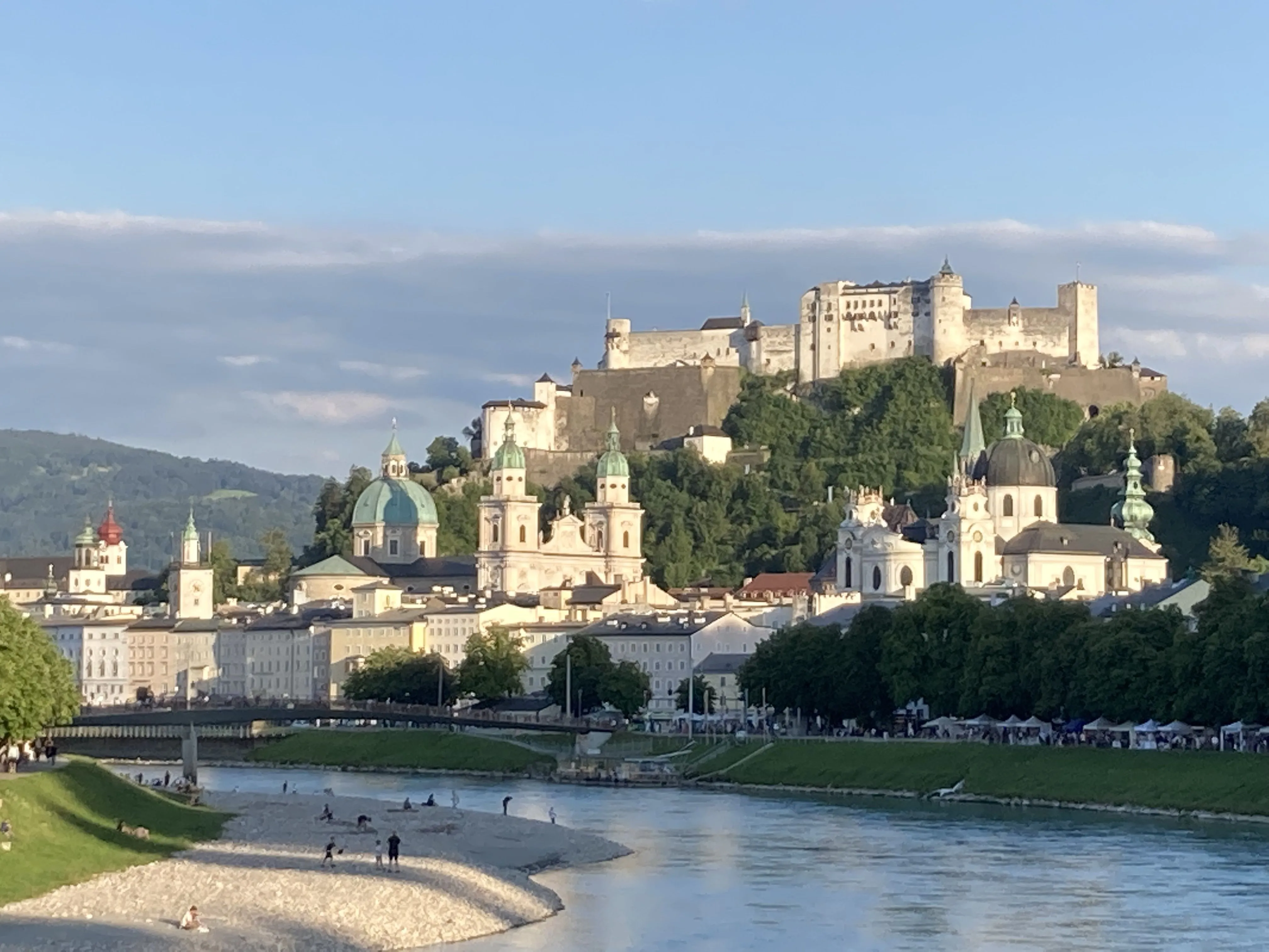 Amplia vista panorámica de la fortaleza de Hohensalzburg situada en una colina boscosa sobre Salzburgo, con el río Salzach, la ribera y las cúpulas de la catedral barroca visibles en primer plano.