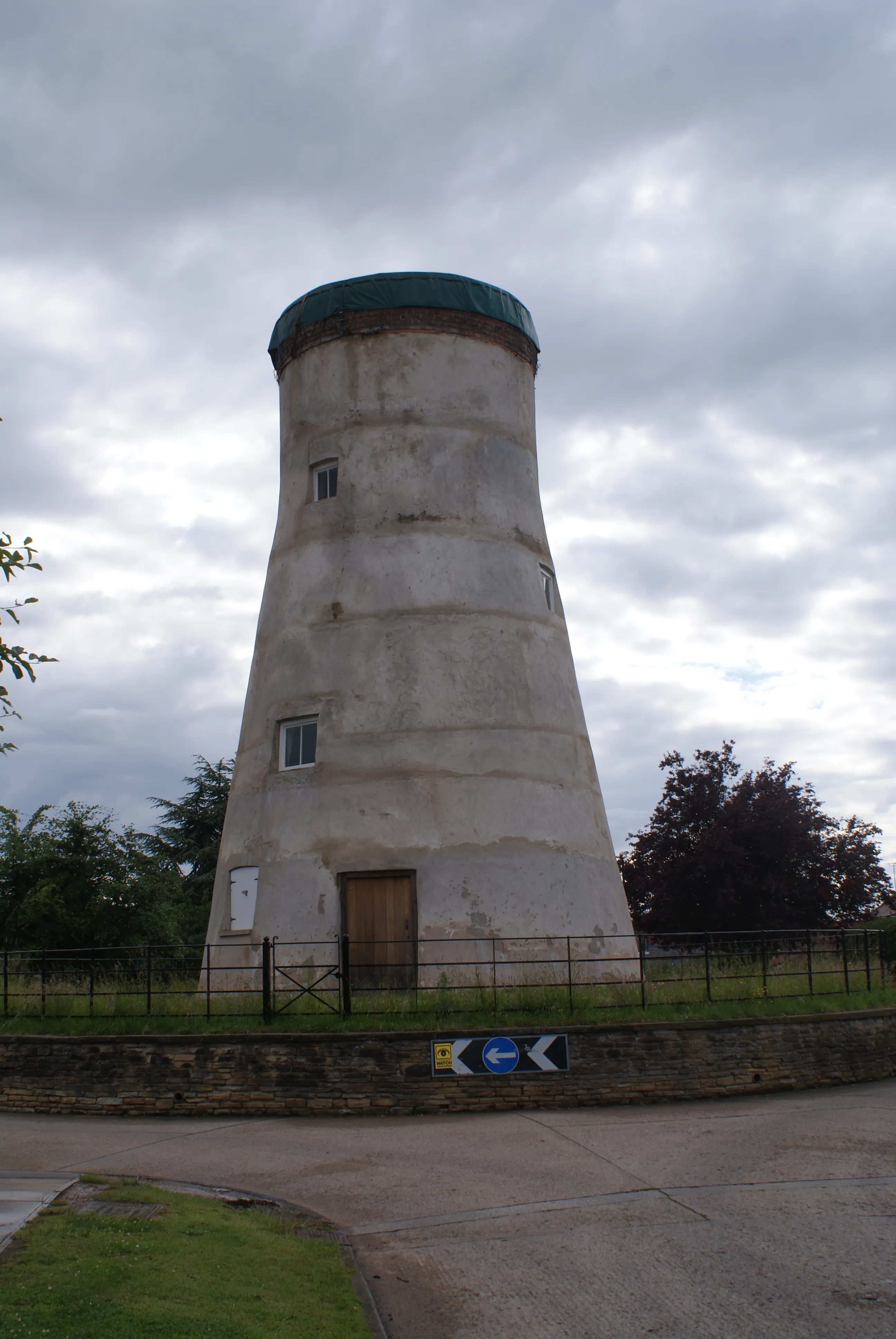 Alta torre de molino blanca y cónica con una cúpula verde, ventanas pequeñas y una puerta de madera en la base, rodeada por una valla de metal negro bajo un cielo nublado.