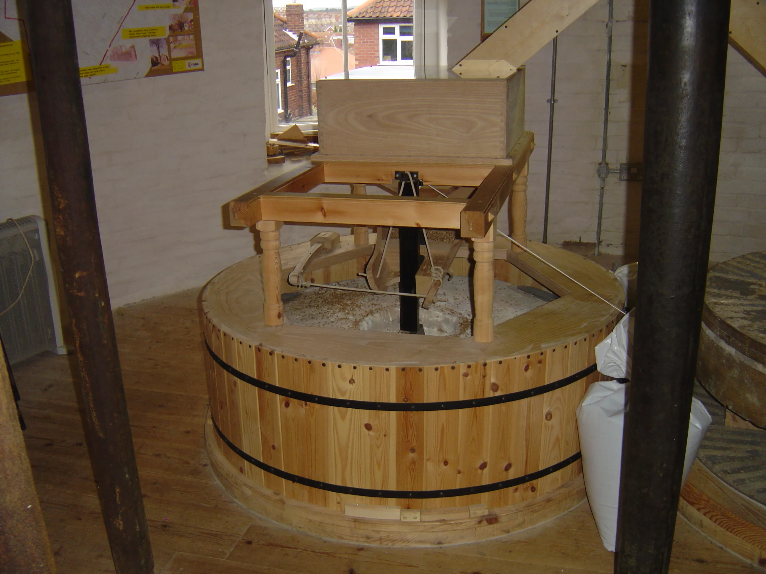 Maquinaria de molino de madera dentro de Holgate Windmill que muestra una gran tina circular de madera con una estructura de marco de madera encima, con una ventana que revela edificios de ladrillo en el exterior.