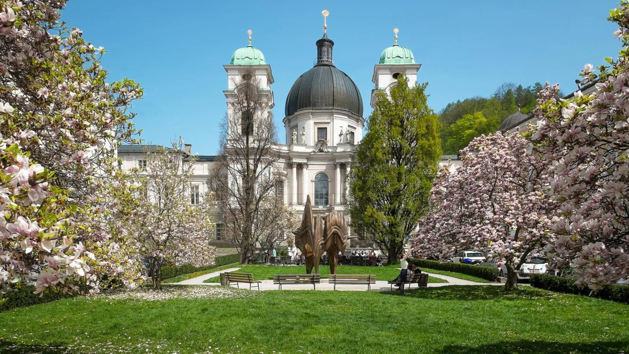 La Iglesia de la Santísima Trinidad en Salzburgo con su cúpula central y torres gemelas, enmarcada por cerezos en flor de color rosa y blanco en un jardín primaveral con una escultura de bronce sobre un césped verde.