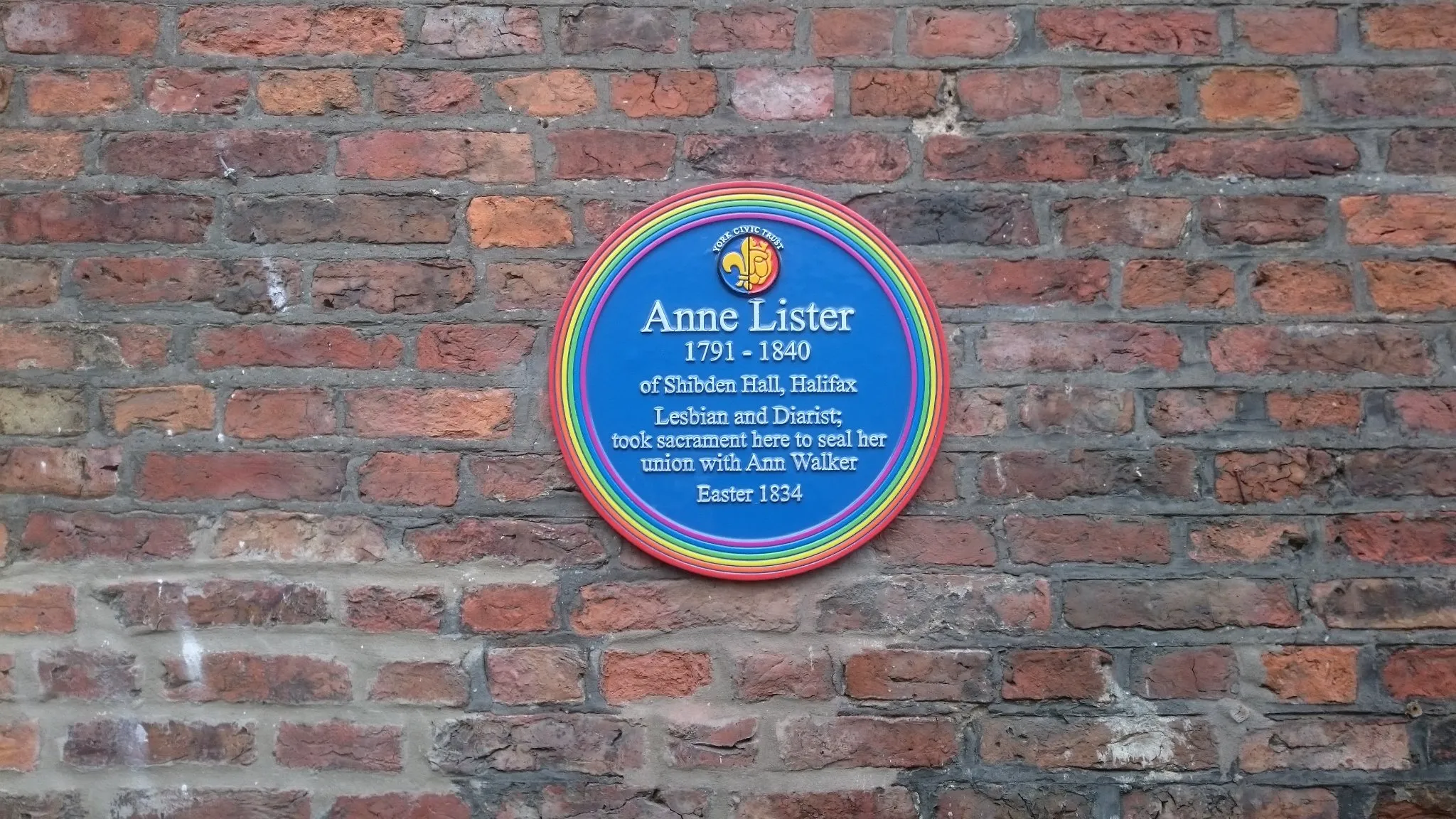 Placa conmemorativa circular azul dedicada a Anne Lister (1791–1840) montada en una pared de ladrillo rojo, que señala que vivió y murió en la Holy Trinity Church y hace referencia a su unión con Ann Walker.