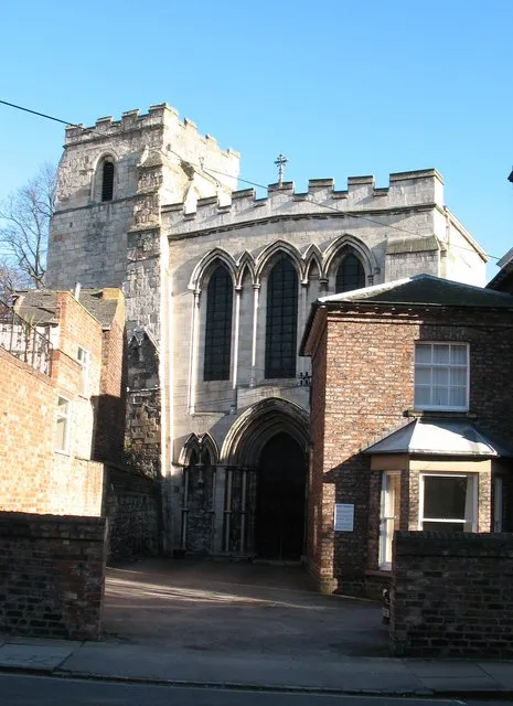 Stenen kerk exterieur met een vierkante toren, gotische spitsboogvensters en een spitsbogige ingang, geflankeerd door bakstenen gebouwen onder een strakblauwe hemel