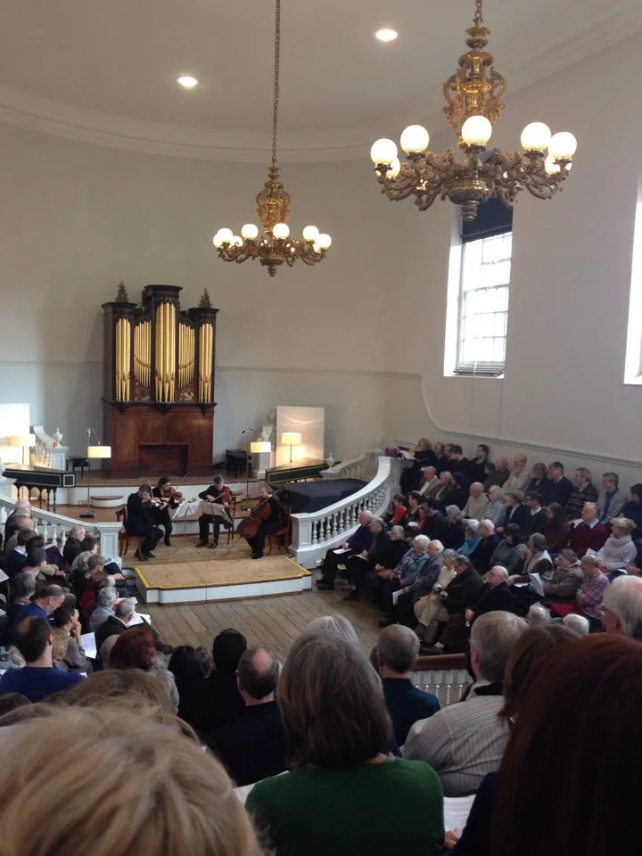 Interieur van de Holywell Music Room met een strijkkwartet dat optreedt op een verhoogd podium, met een publiek gezeten in een hoefijzervormige opstelling, een groot pijporgel aan de achterwand en sierlijke gouden kroonluchters die aan het plafond hangen.