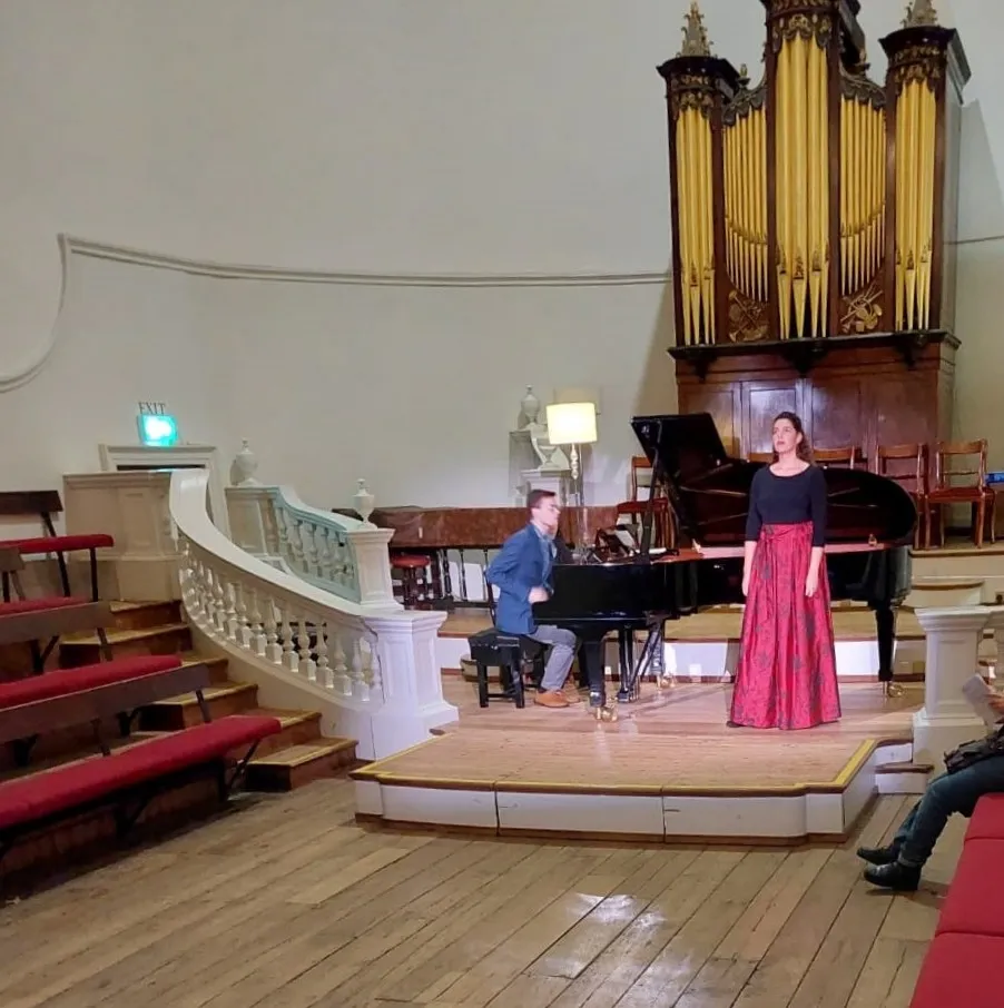 Interieur van de Holywell Music Room met een optreden op een houten podium met een vleugel, een groot sierlijk pijporgel en een gebogen wit balkon met rode stoelen.