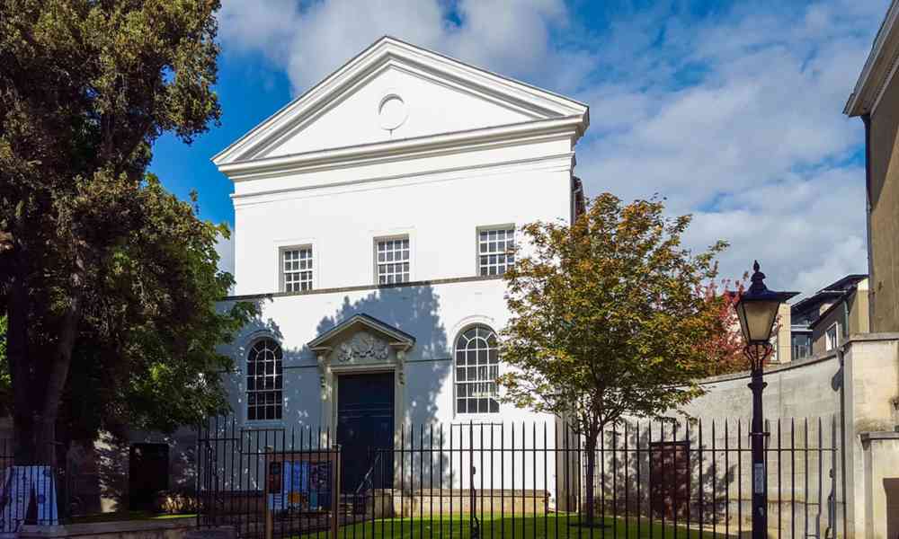 Wit klassiek gebouw met een driehoekig fronton, boogvensters, donkere toegangsdeur en zwart ijzeren hekwerk onder een blauwe lucht met bomen