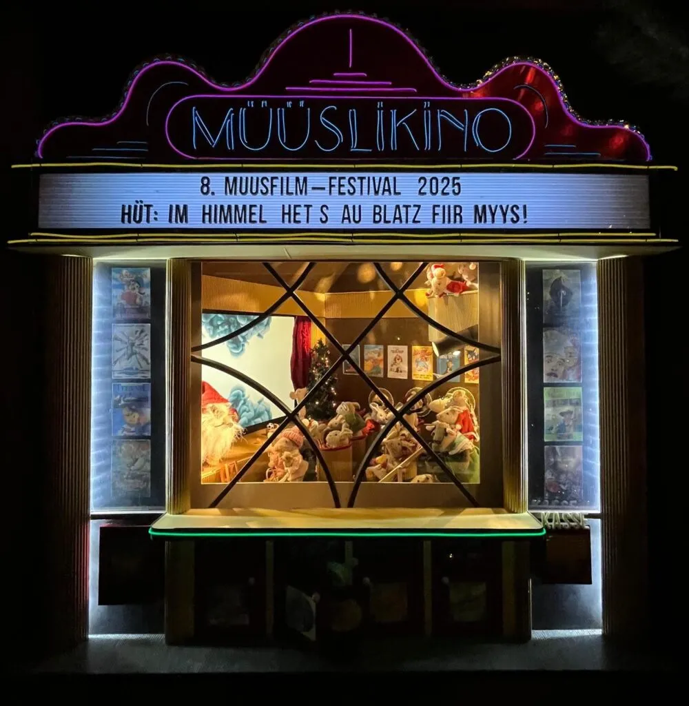 Fachada de cine en miniatura con un letrero de neón rosa que dice MÜÜSLIKINO y una marquesina que anuncia el 8.º Muusfilm Festival 2025, exhibida en una ventana iluminada por la noche con una escena interior brillante visible a través de un cristal con cuarterones.