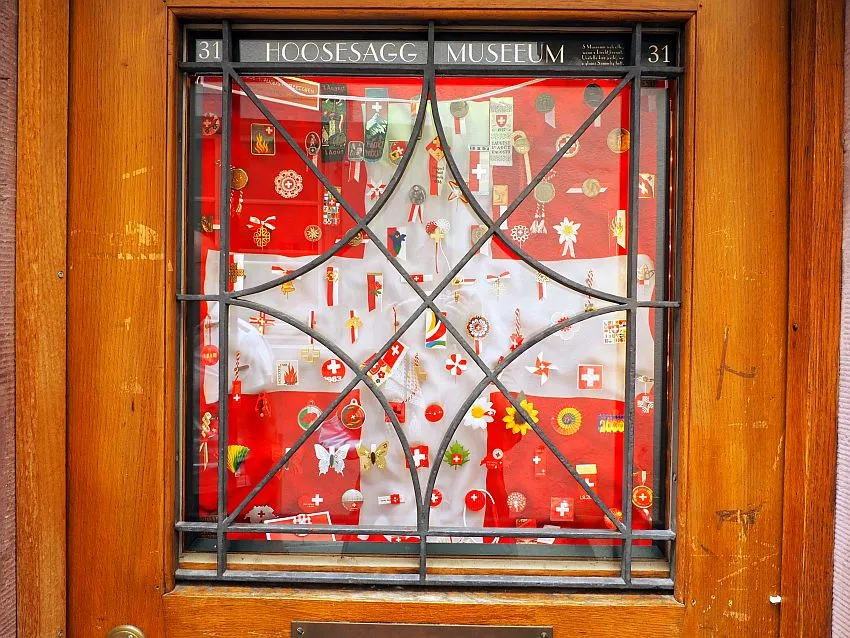 Escaparate del Hoosesaggmuseum con una exposición en miniatura de color rojo y blanco detrás de una reja de hierro decorativa, con el letrero del nombre del museo arriba