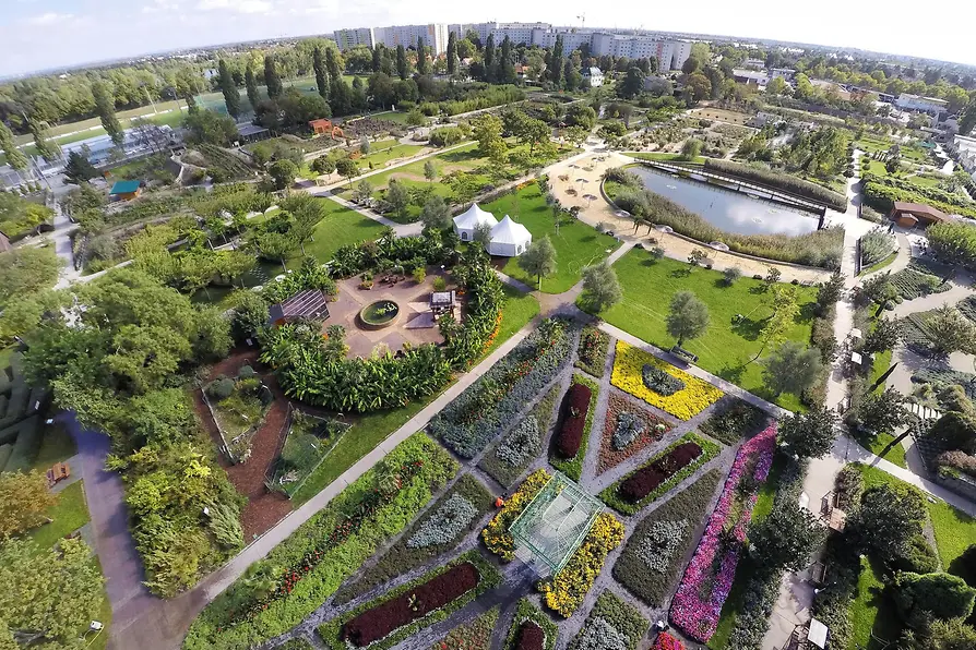 Aerial view of a large garden with colorful flower beds, pathways, a pond, and a white tent in the center.