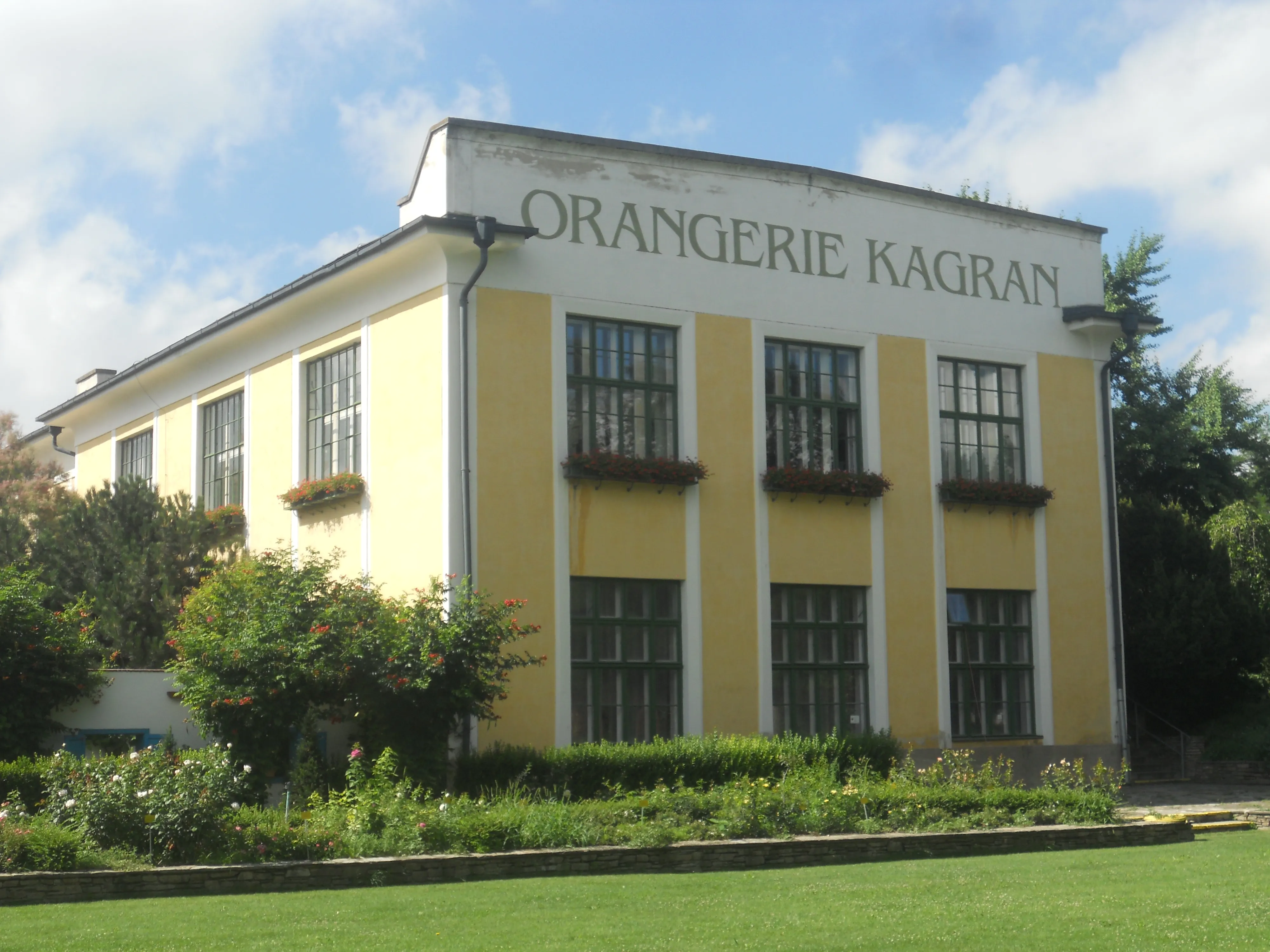yellow building with white trim and 'ORANGERIE KAGRAN' sign, surrounded by greenery and lawn
