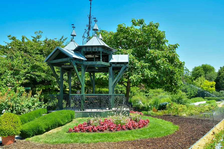 A green metal gazebo with ornate details in a landscaped garden with red flowers and green shrubs under a blue sky