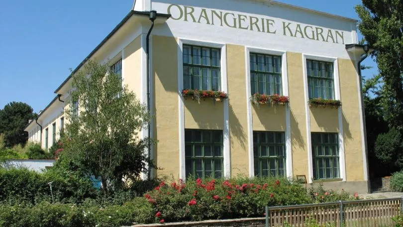 yellow building with green windows and flower boxes, surrounded by greenery and red flowers