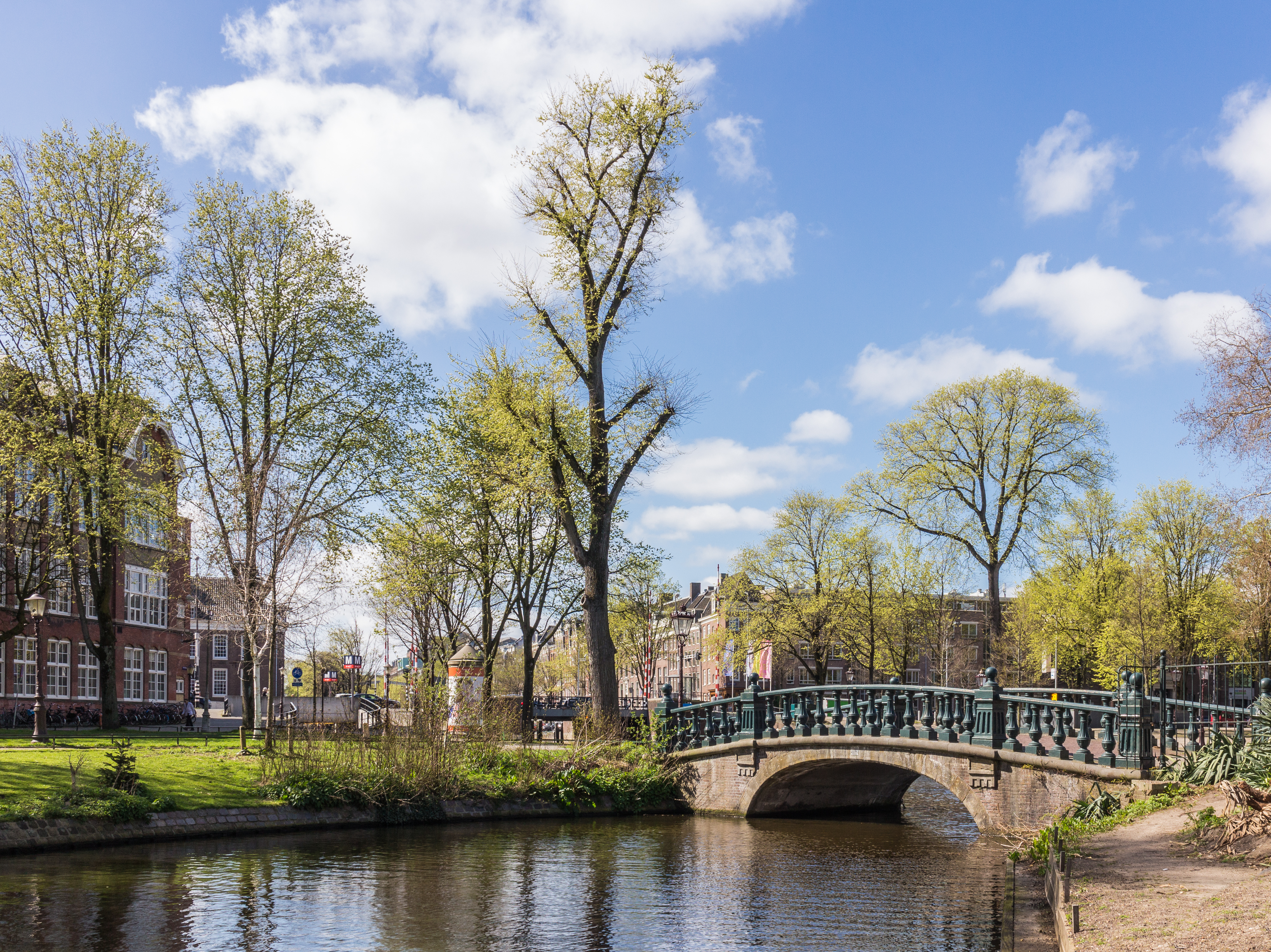 Zielony, łukowy most nad kanałem w Hortus Botanicus Amsterdam z drzewami i budynkami w tle pod błękitnym niebem
