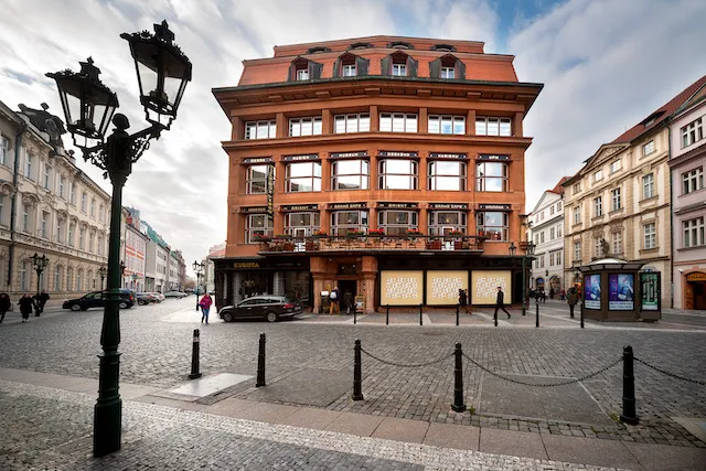 Czerwony budynek z cegły z geometrycznymi oknami i zakrzywioną fasadą, z czarną latarnią uliczną na pierwszym planie na brukowanej ulicy.