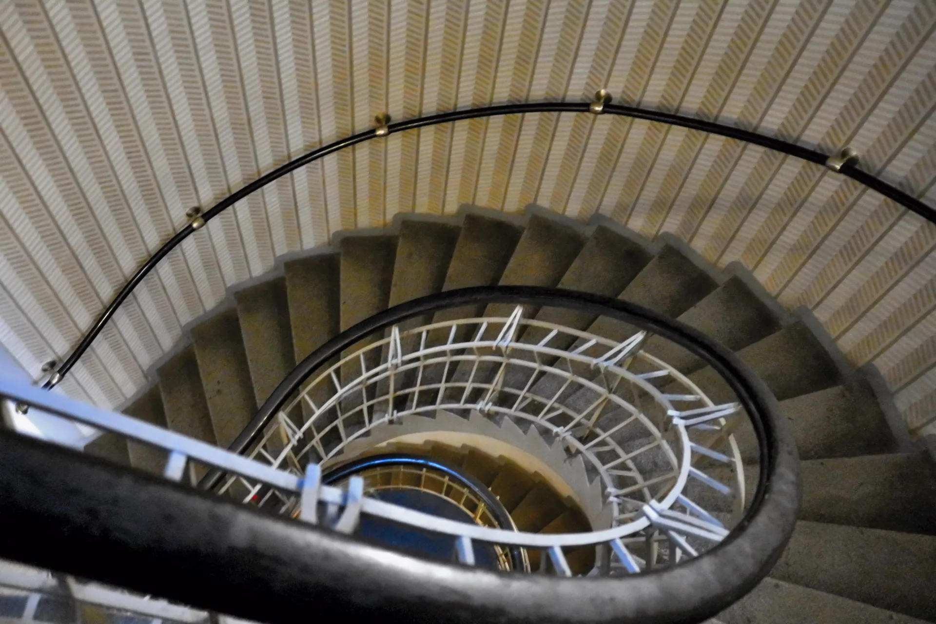 Kręcone schody z czarną metalową balustradą i białym metalowym elementem wspierającym, widziane z góry na tle teksturowanej beżowej ściany.