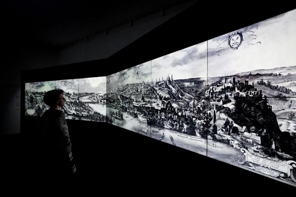 Silhouet van een persoon die een panoramische historische illustratie van een stad bekijkt op een gebogen muur in een donkere kamer