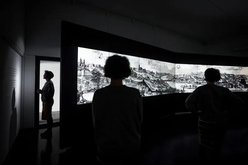 Silhouetten van drie mensen die een groot zwart-wit panoramisch kunstwerk bekijken in een donkere galerie.