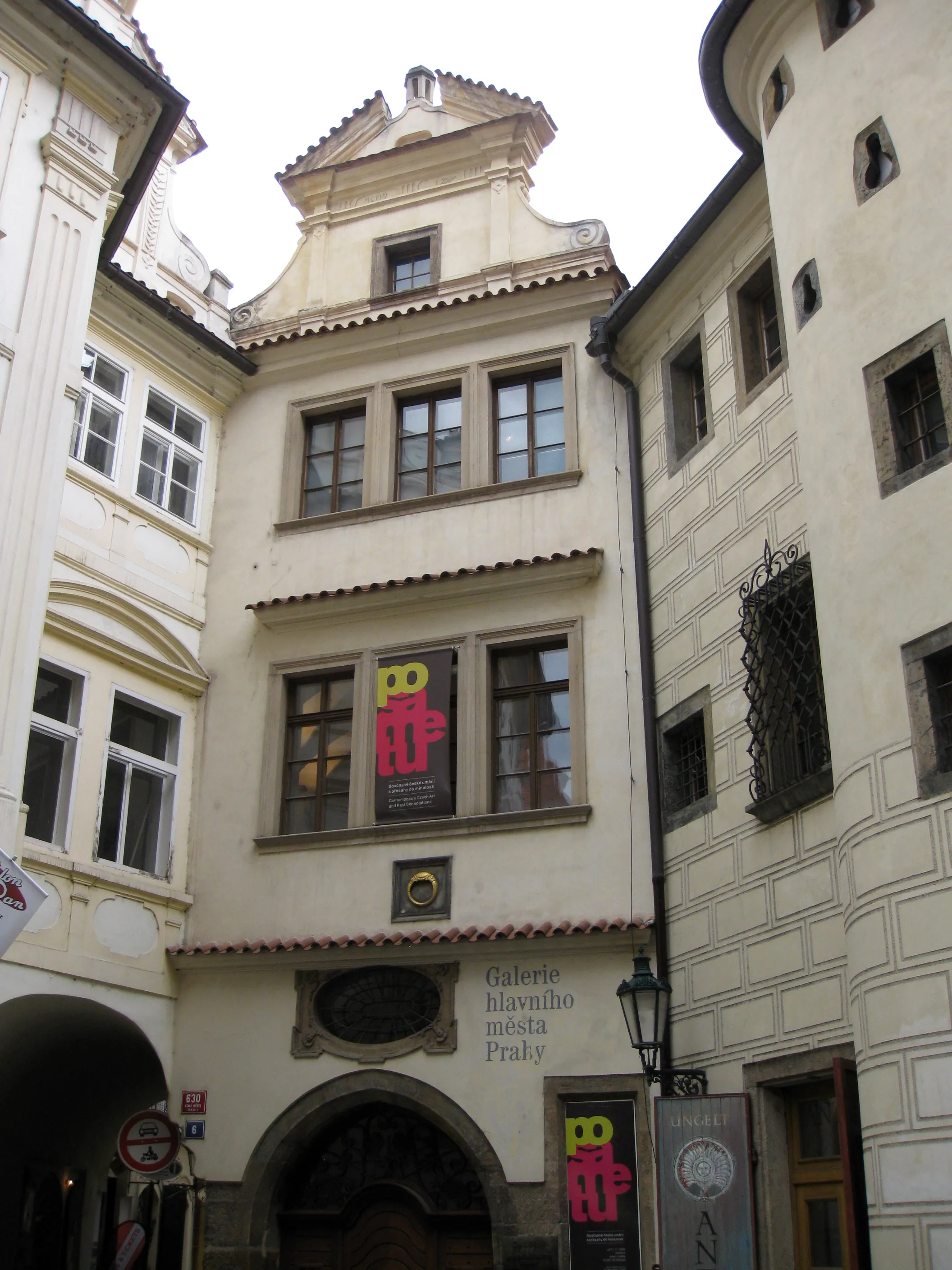 Un edificio estrecho y histórico con paredes de color crema y ventanas arqueadas, que presenta un letrero de la Galerie hlavního města Prahy y coloridos carteles de exposiciones en los escaparates.