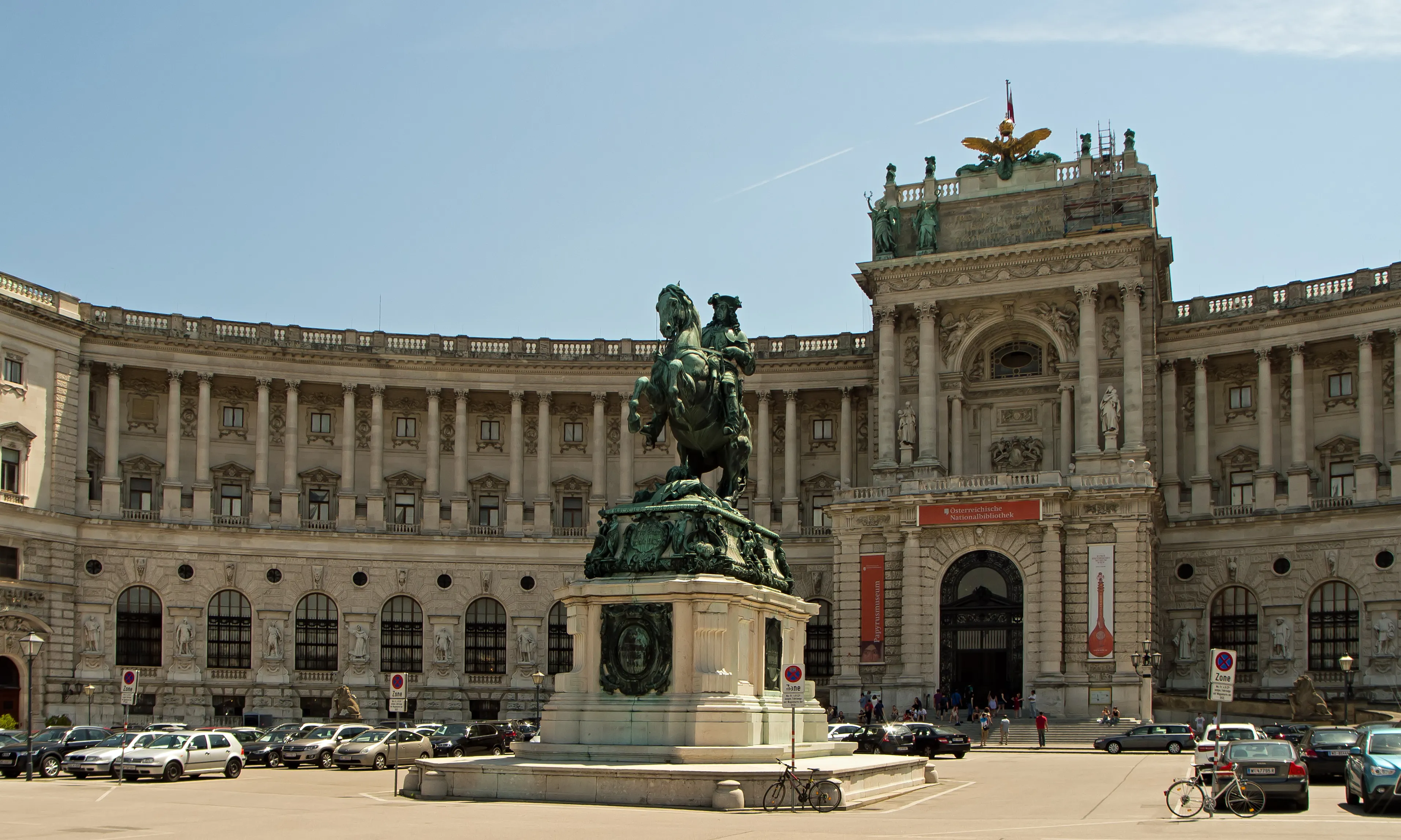 A large classical building with a central entrance and a green equestrian statue in front, under a clear blue sky
