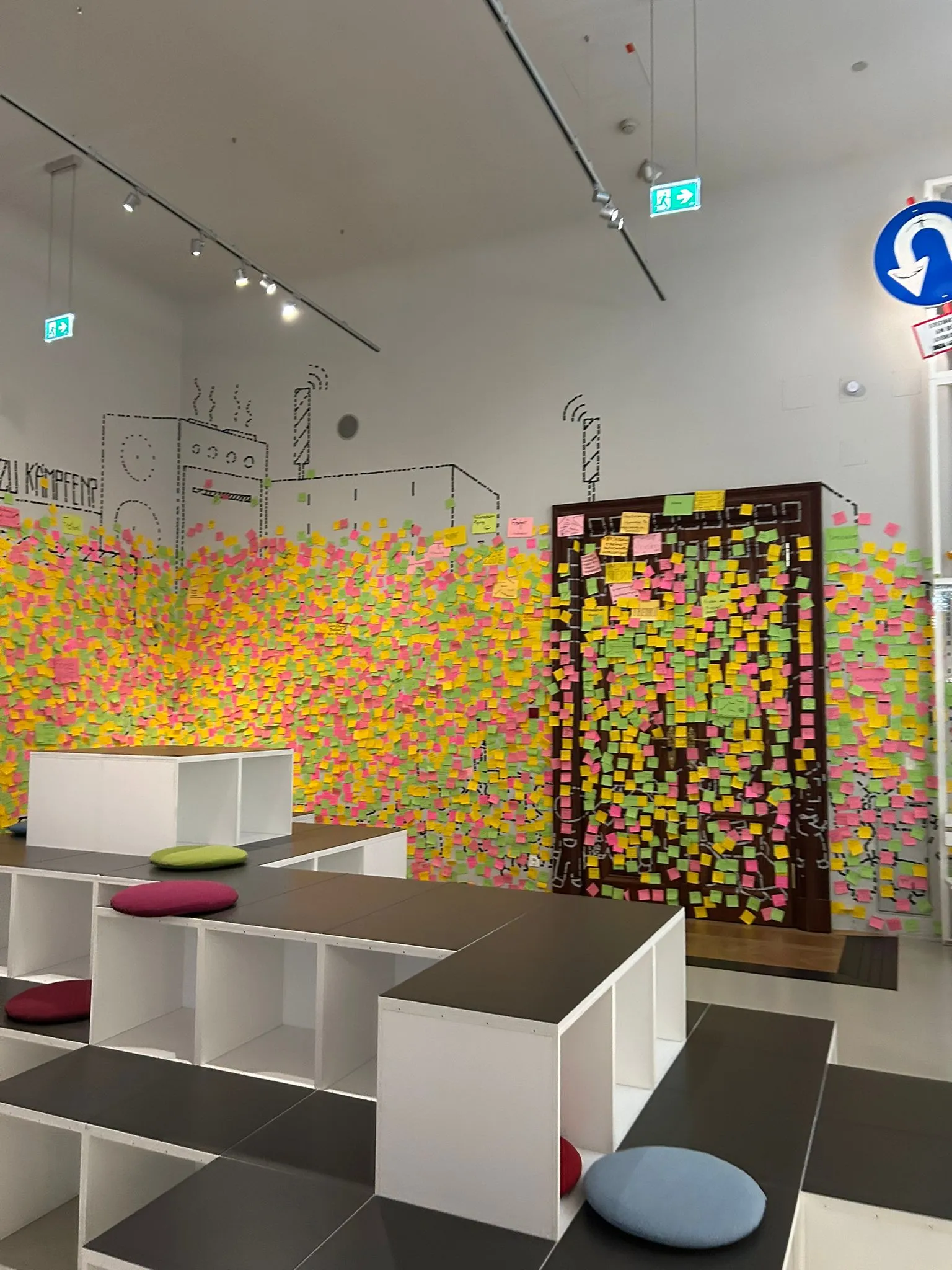 A modern exhibition space with a wall covered in colorful sticky notes and a wooden board filled with notes, with white modular seating and cushions in the foreground.