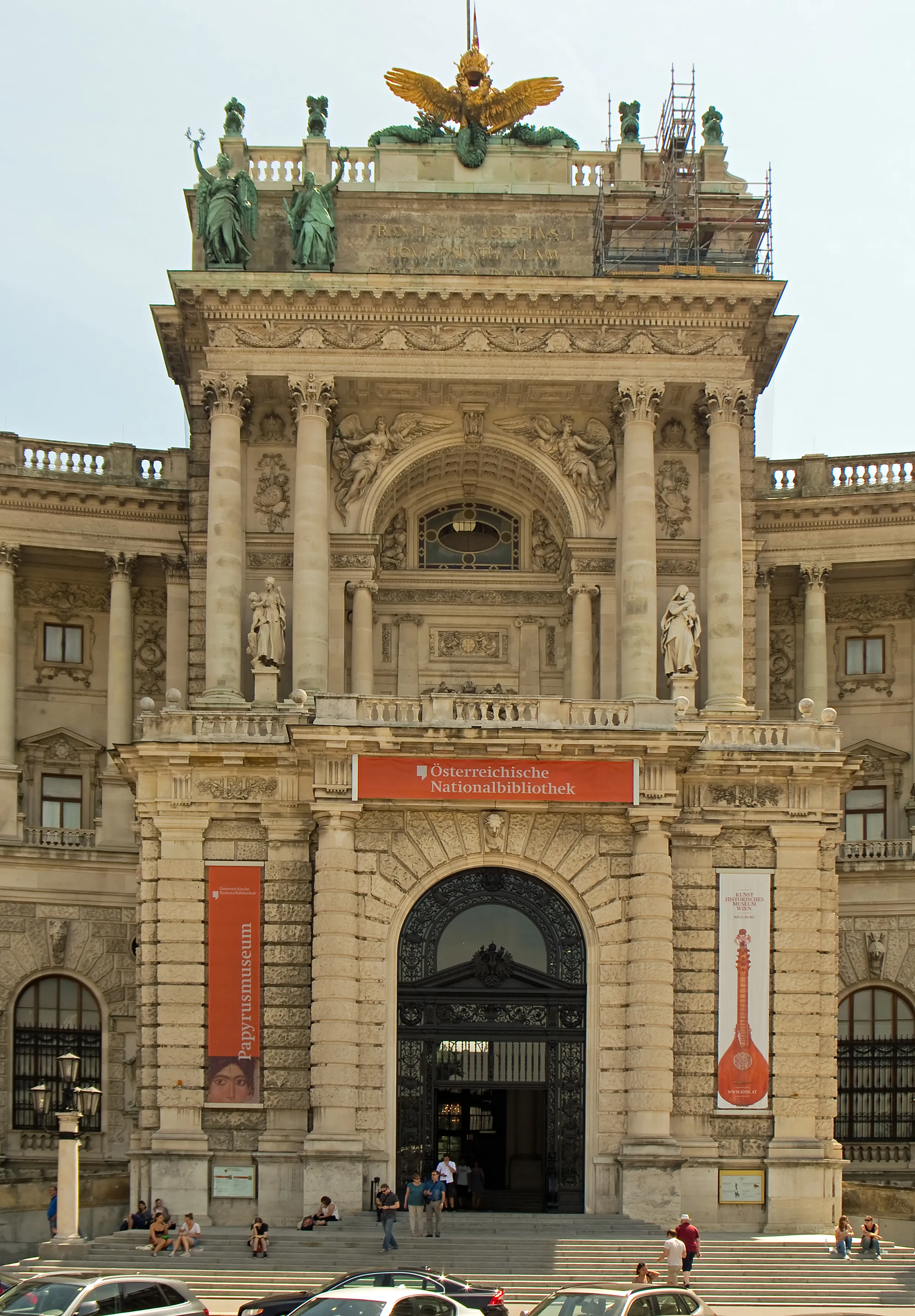 The ornate stone facade of the House of Austrian History, featuring columns, statues, a golden eagle, and a red banner with text, with people and cars in the foreground.