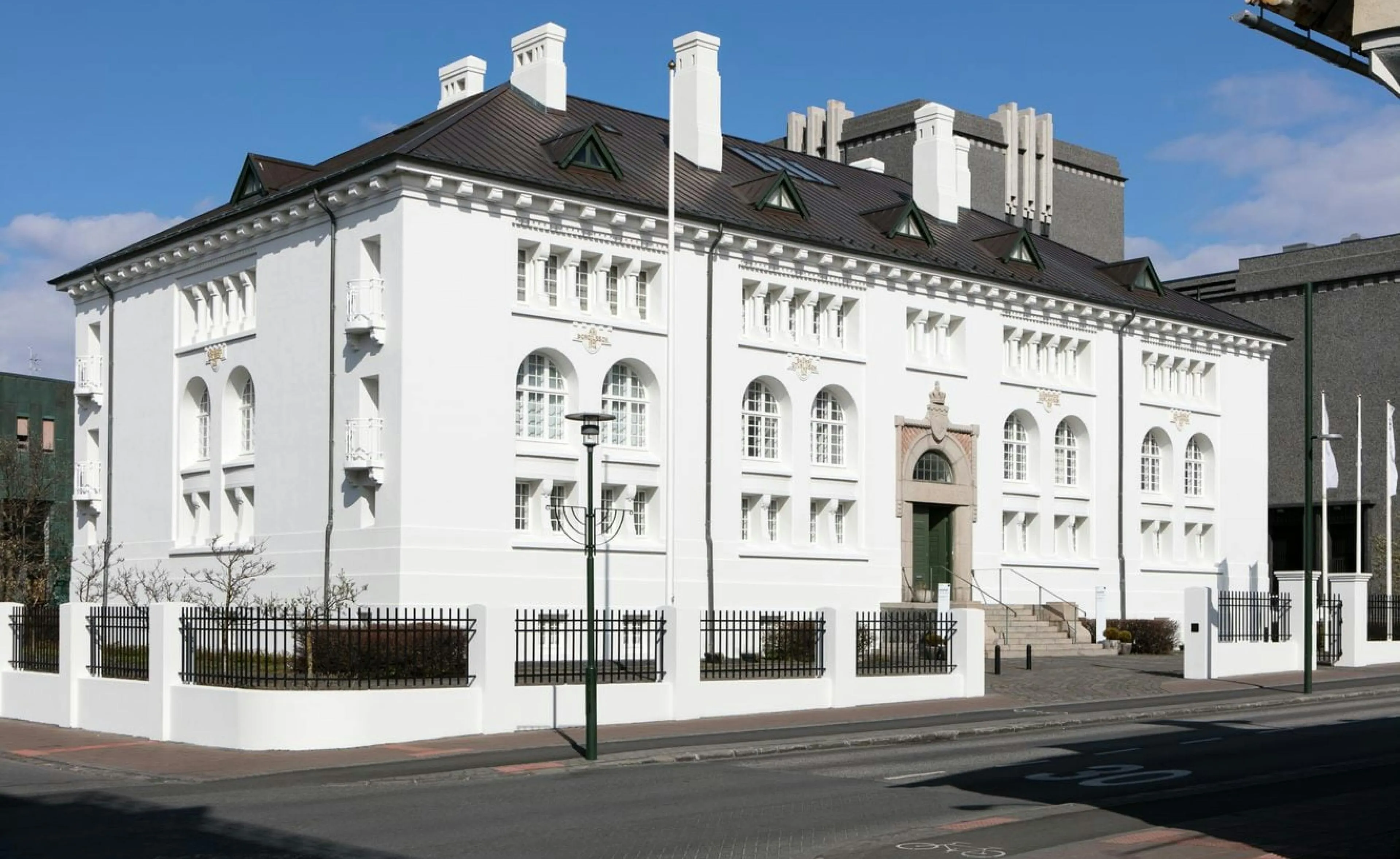 Biały zabytkowy budynek z ciemnym dachem, łukowymi oknami i zielonymi drzwiami wejściowymi, otoczony niskim białym murem z czarnym żelaznym ogrodzeniem