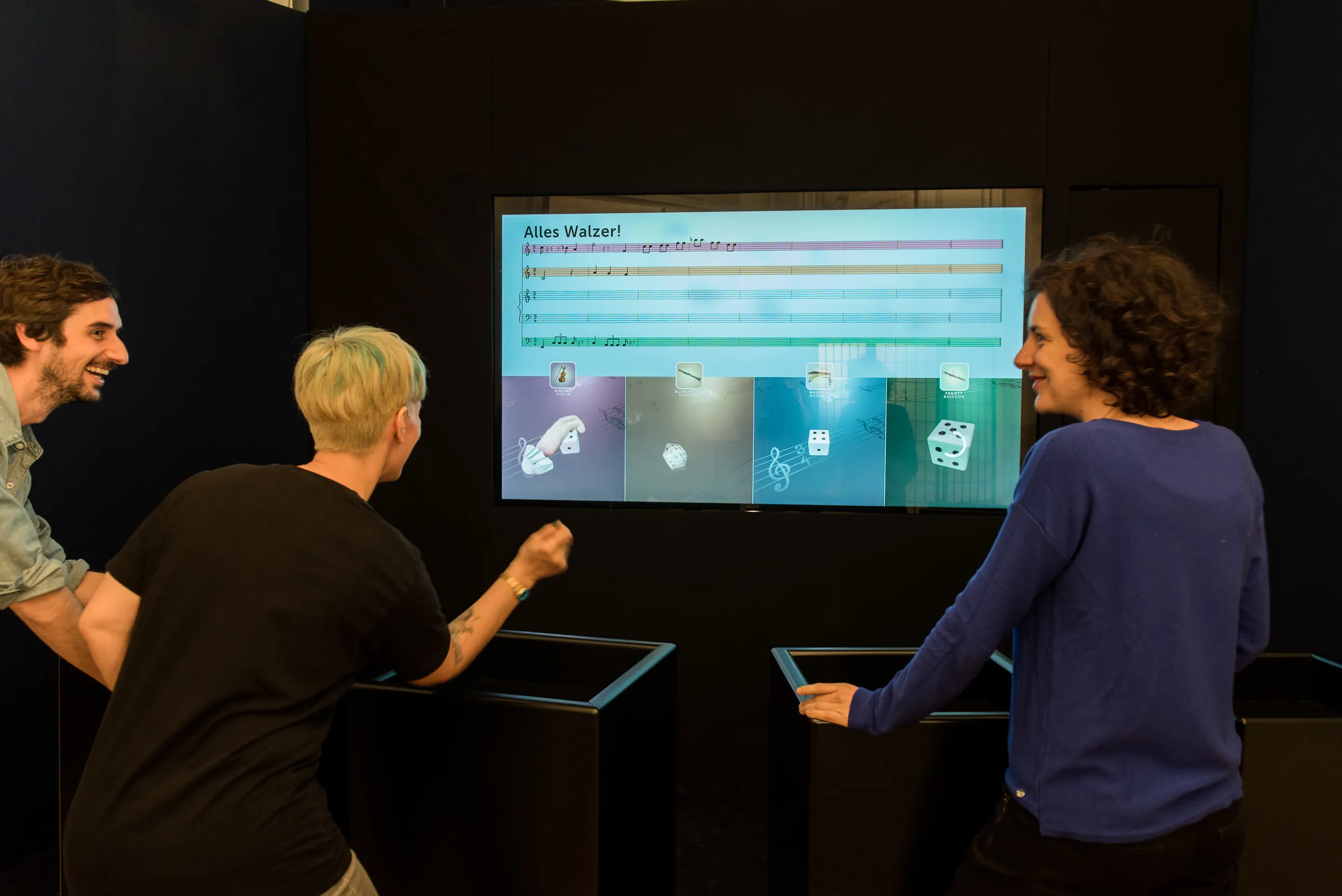 Tres personas interactuando con una exposición de música digital en una gran pantalla que muestra íconos y texto coloridos
