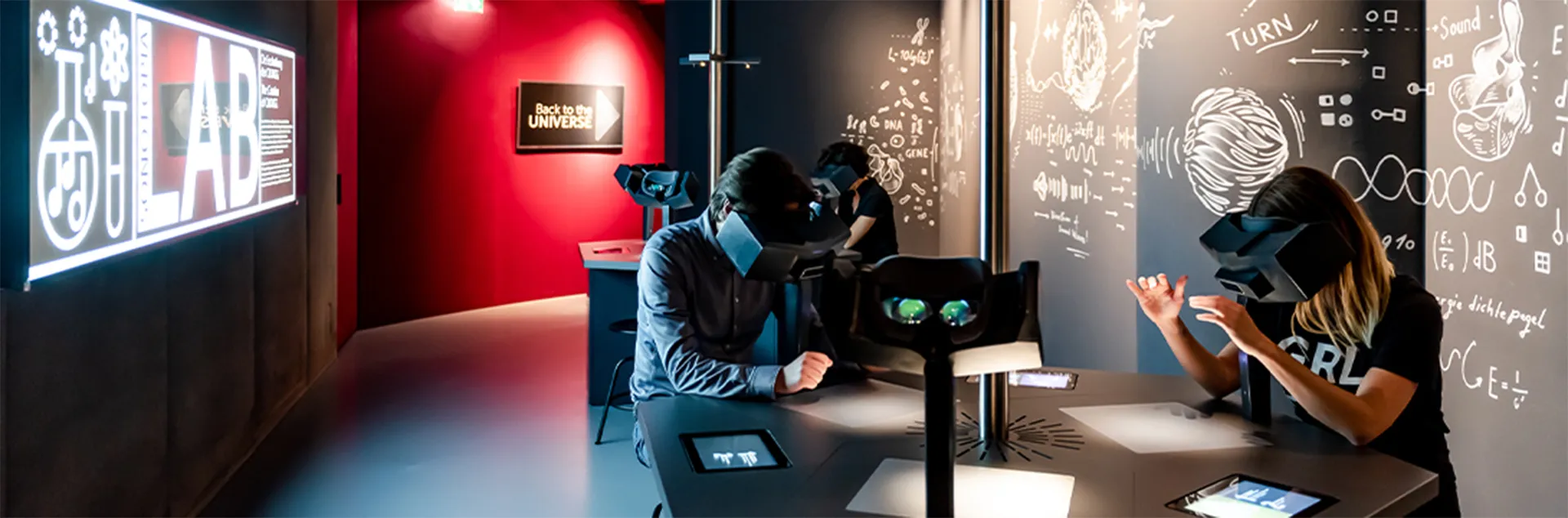 Dos personas con cascos de RV en una mesa con equipo de sonido interactivo en una habitación oscura con paredes rojas y un letrero brillante de 'LAB'.