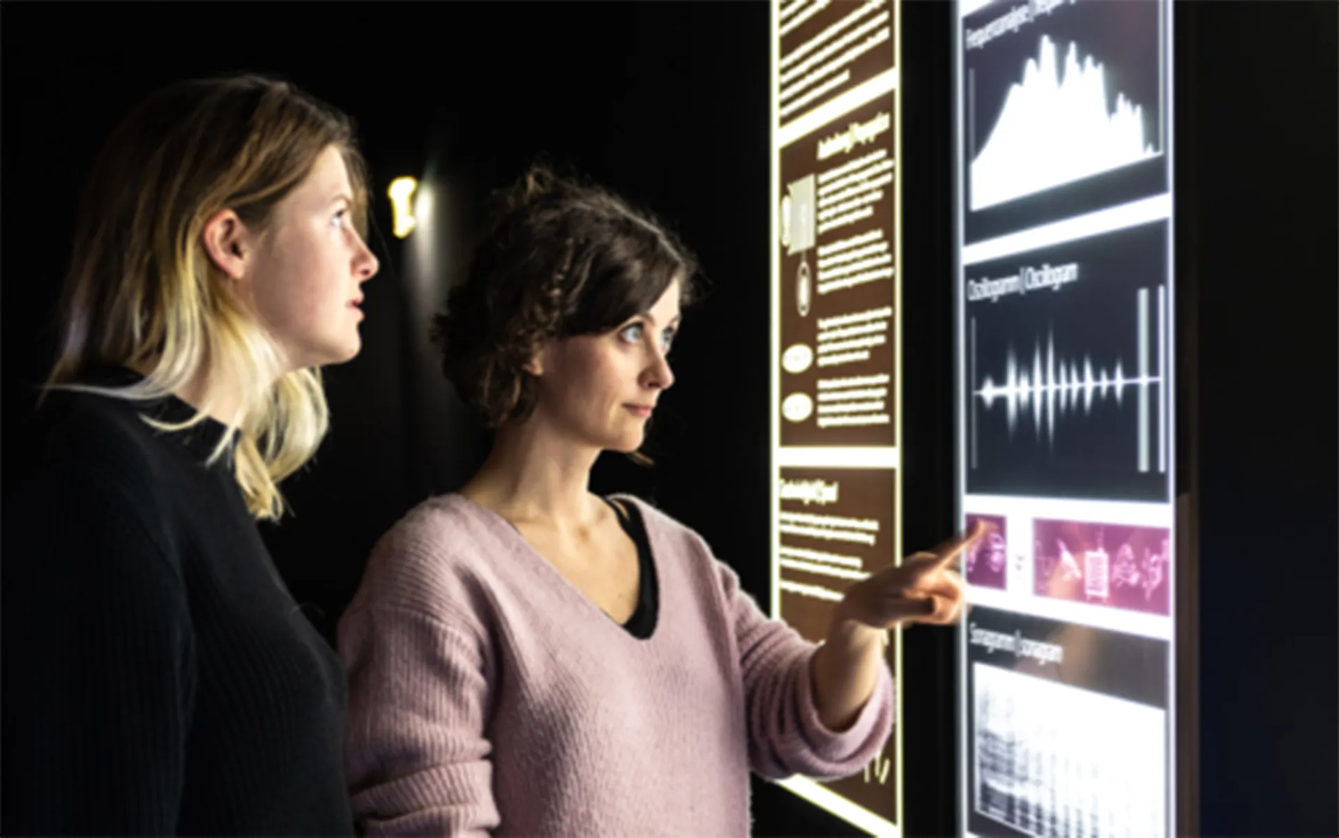 Dos mujeres mirando una pantalla interactiva que muestra ondas sonoras y datos.