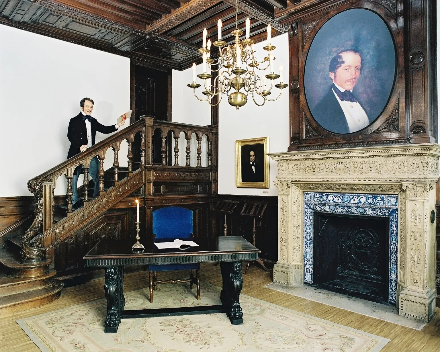 Habitación interior ornamentada con una escalera de madera, candelabro, chimenea y un gran retrato en la pared
