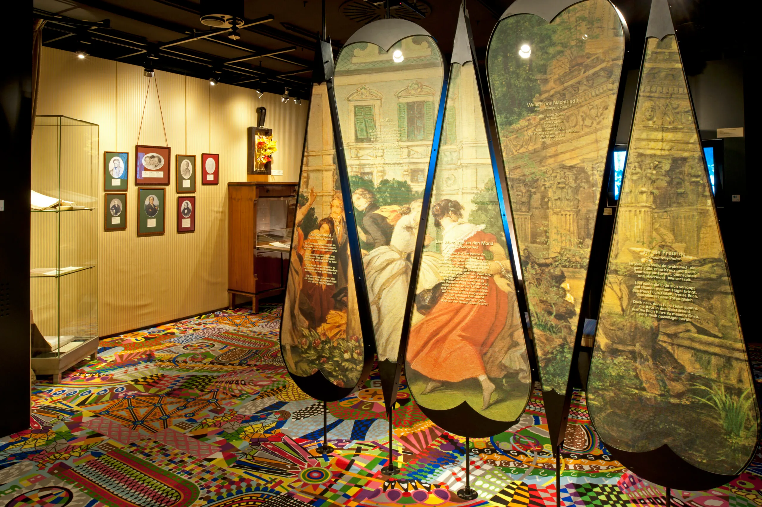 Paneles de exhibición grandes en con forma de lágrima con obras de arte y texto en una galería de museo con un suelo alfombrado y colorido.