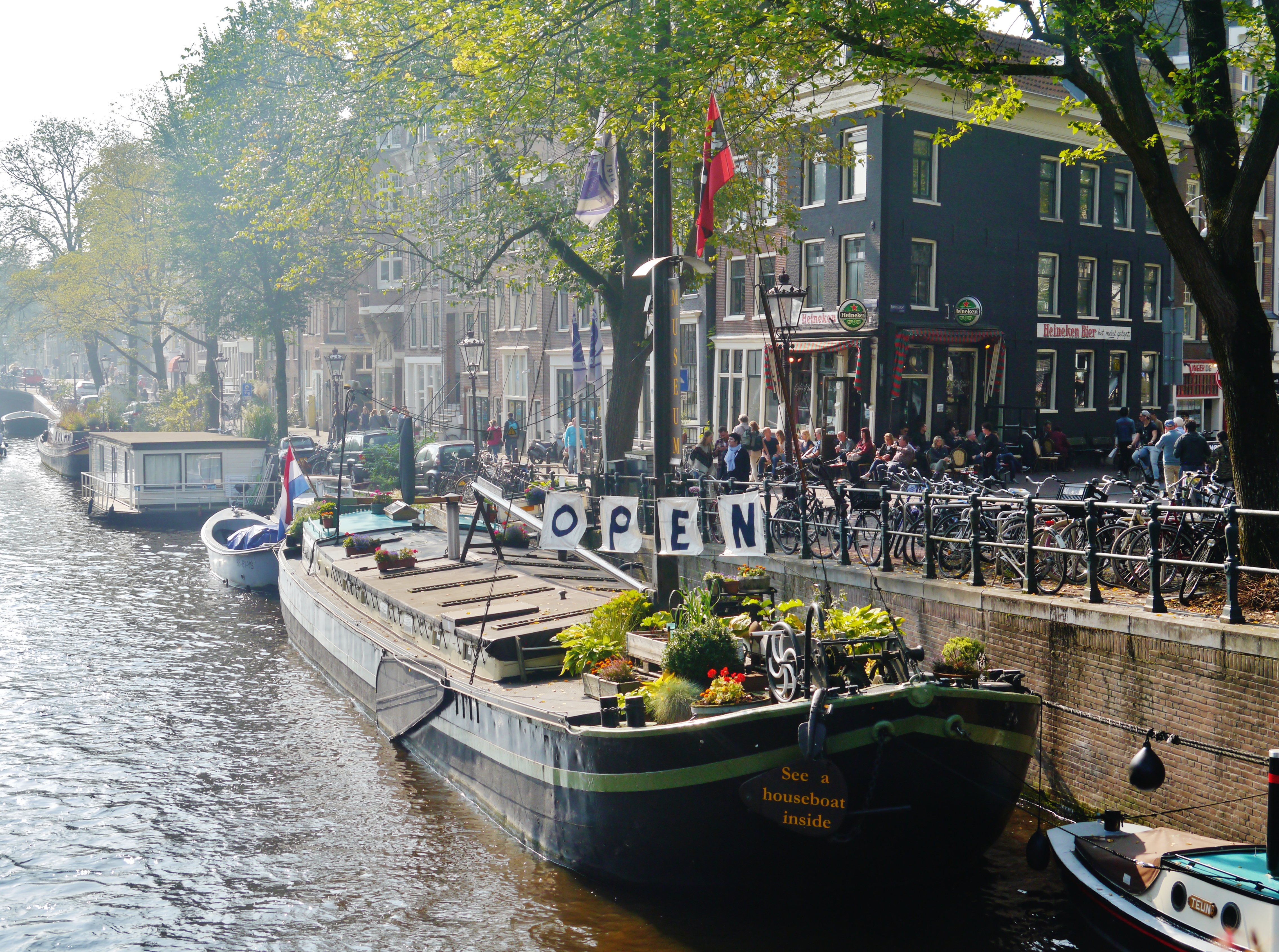 Woonbootmuseum aan een gracht met 'OPEN'-bord, mensen, fietsen en historische gebouwen langs de waterweg in Amsterdam