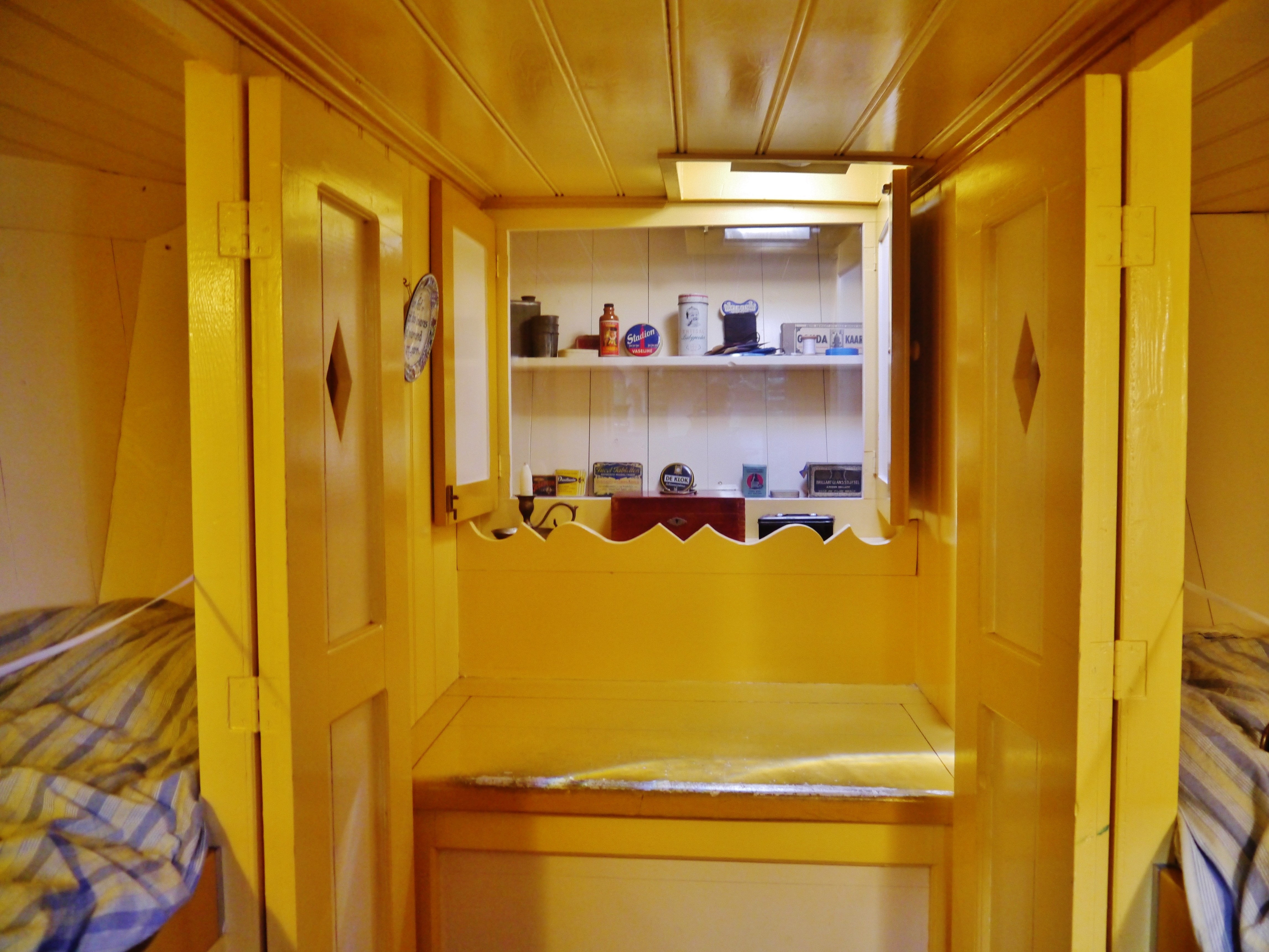 Yellow-painted interior of a houseboat with wooden doors, shelves holding kitchen items, and striped bedding on beds.