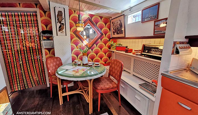 Kleurrijk interieur van een woonboot met een patroon op het behang, een ronde tafel met vier stoelen, een keukenhoek en nautische decoraties op de achtergrond.